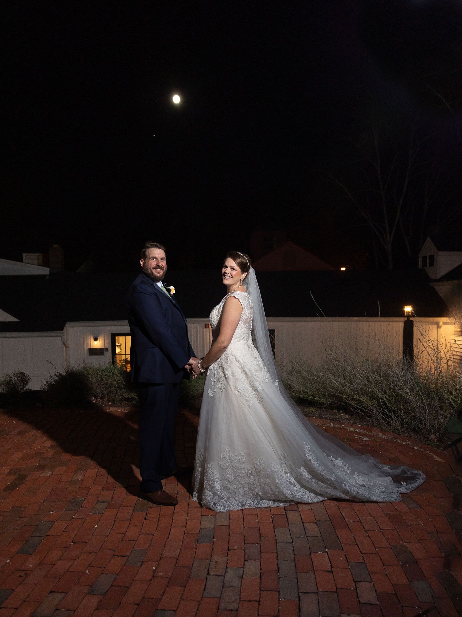 Newlyweds pose on a brick patio at night with a full moon glowing in the dark sky above them.