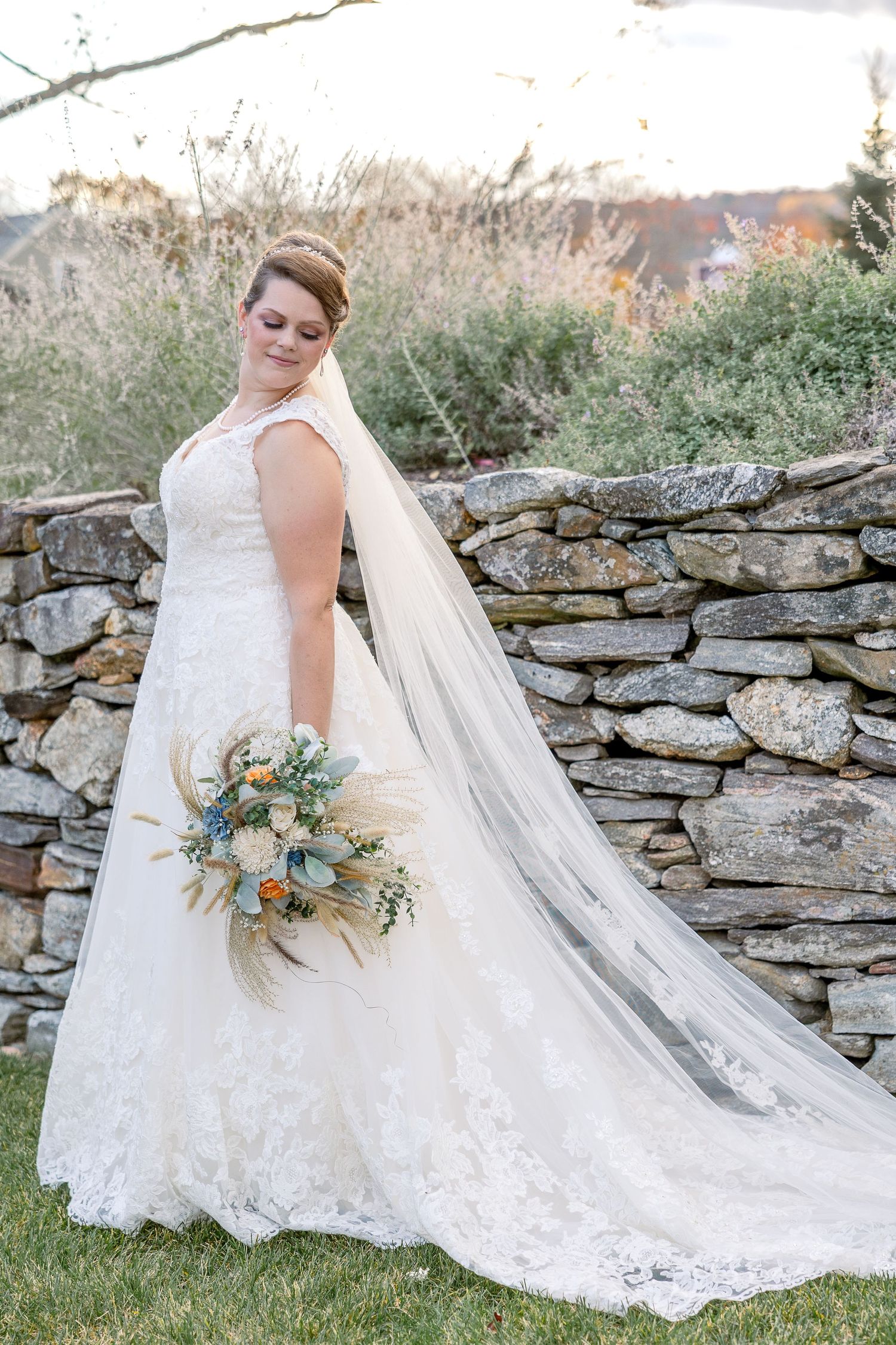 Elegant bride in lace gown with cathedral veil poses by stone wall with floral bouquet.