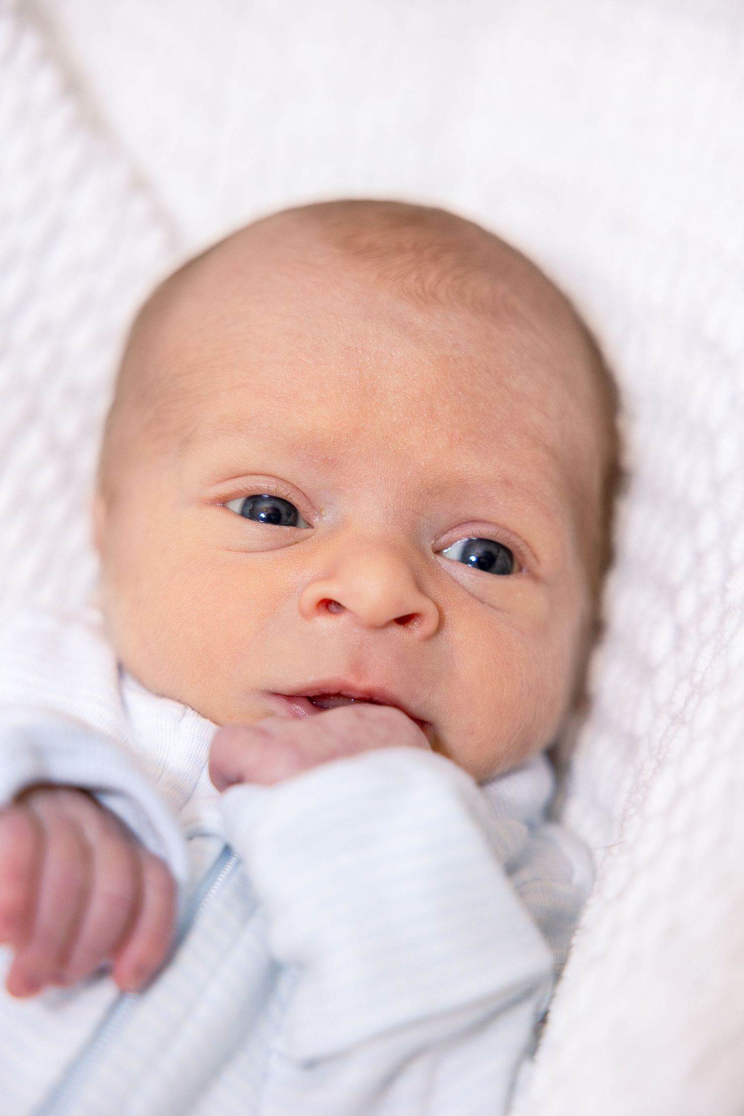 A newborn baby with bright eyes lies swaddled in soft white blankets while making a sweet expression.