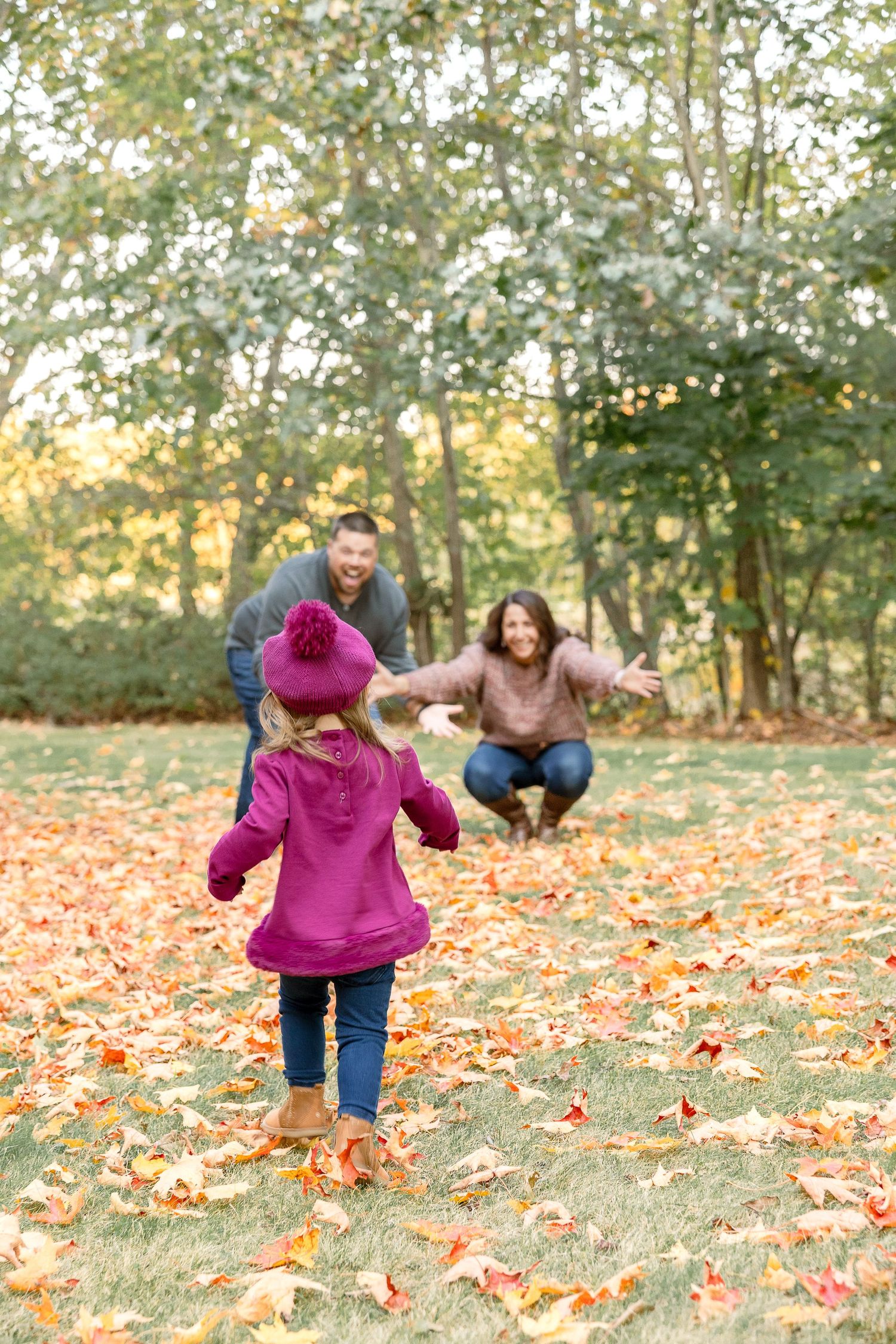 A child in pink coat walks toward parents in leaf-covered autumn ground.