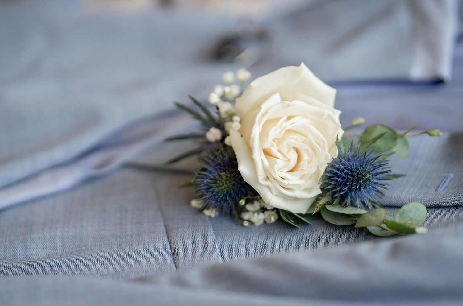 A delicate white rose boutonniere with blue thistle and greenery rests on a light gray fabric background.