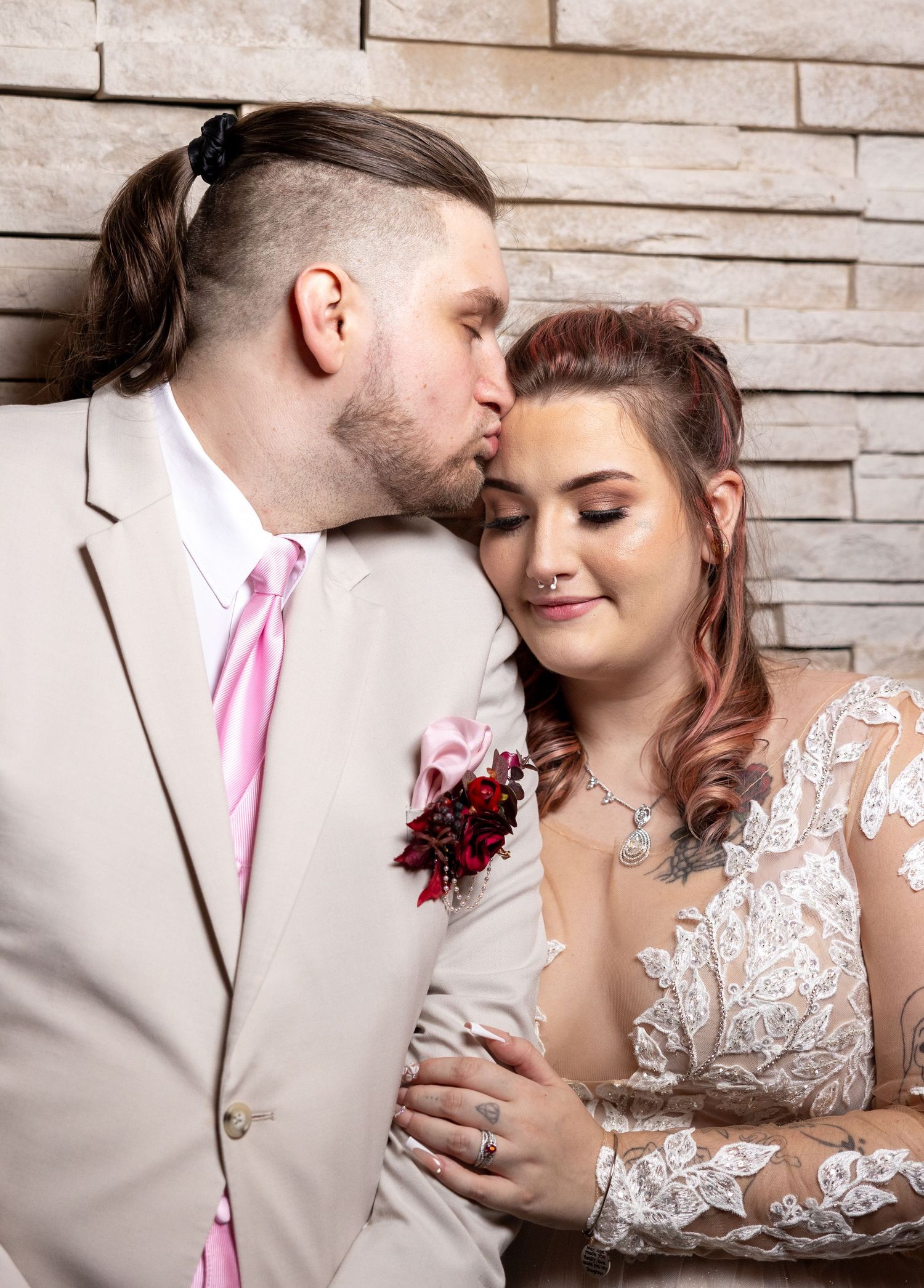 Romantic wedding portrait of couple with beige suit and pink tie embracing against brick wall backdrop.