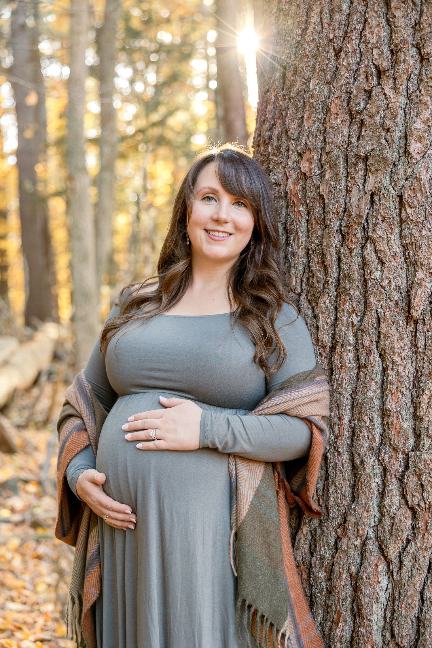 A pregnant person in a grey dress poses by a tree trunk in autumn sunlight with golden forest background.