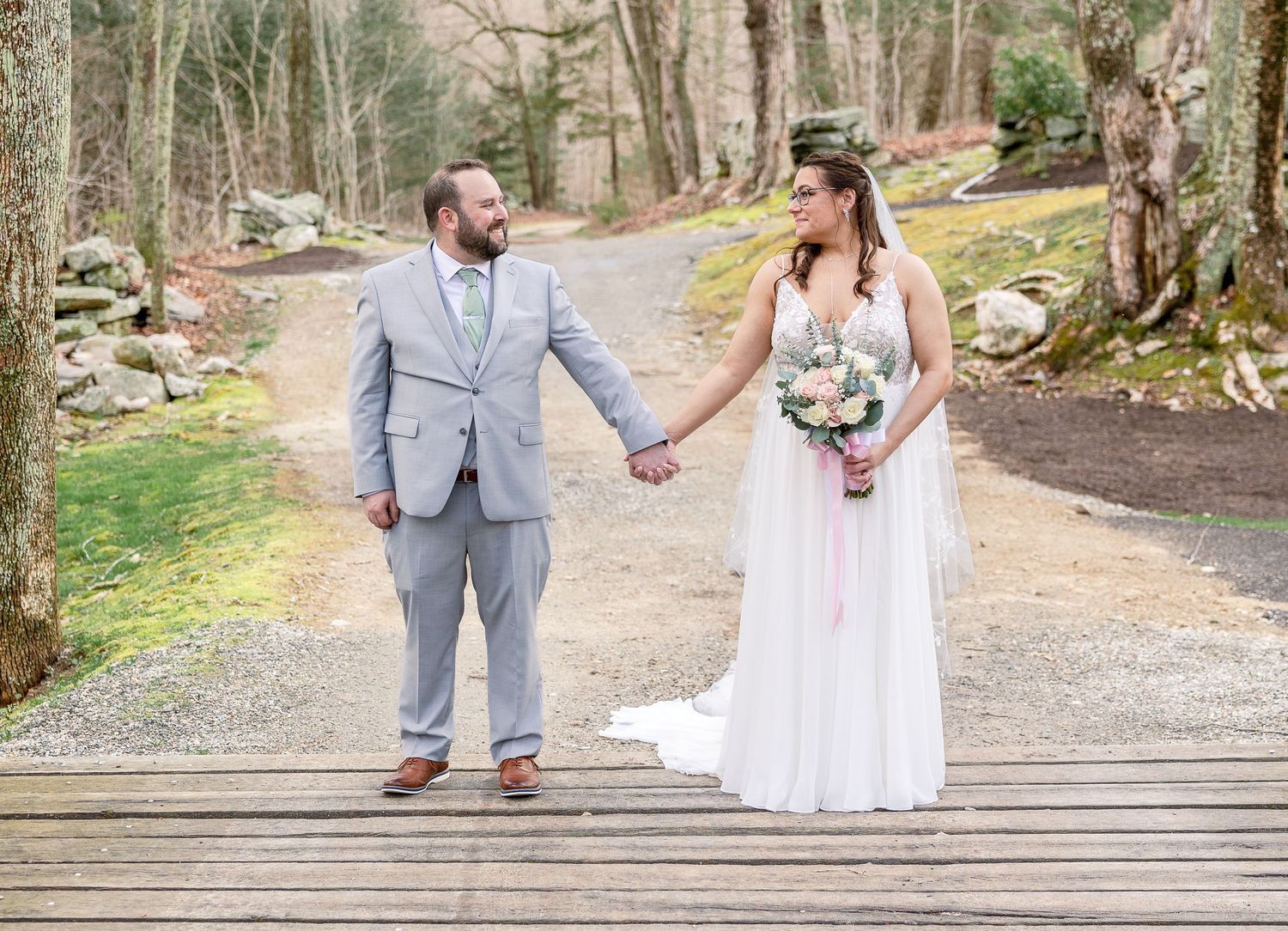A couple in wedding attire holds hands while standing on a wooden bridge along a forest path.