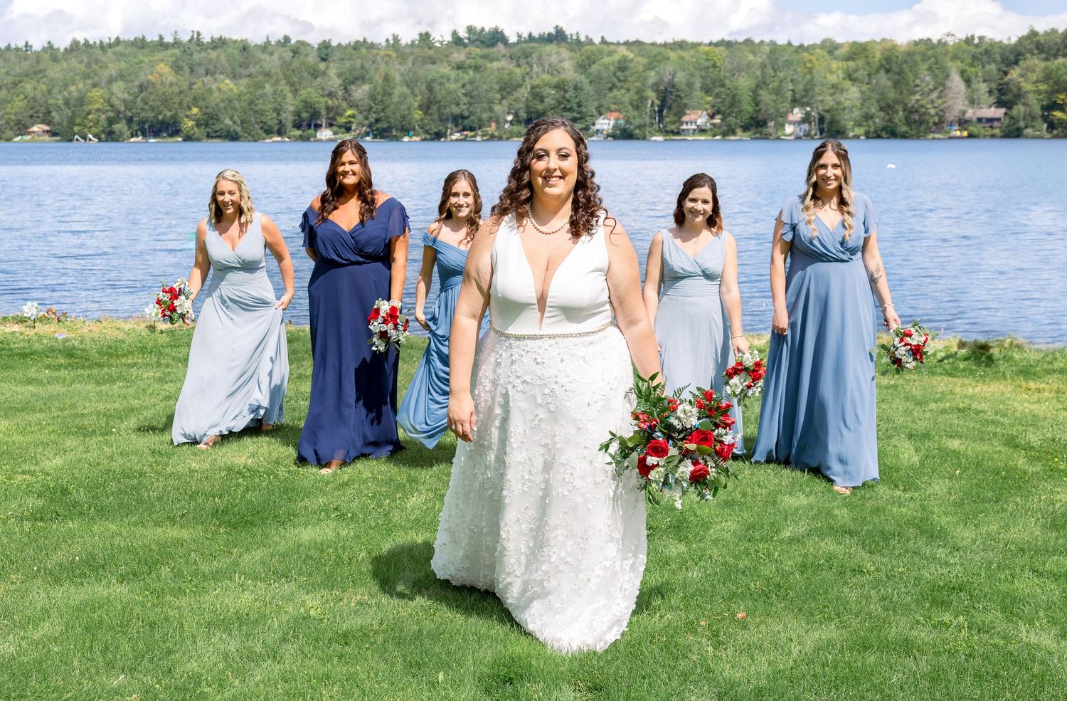 Wedding party in coordinated blue dresses pose on lush grass overlooking scenic lakefront on sunny summer day.