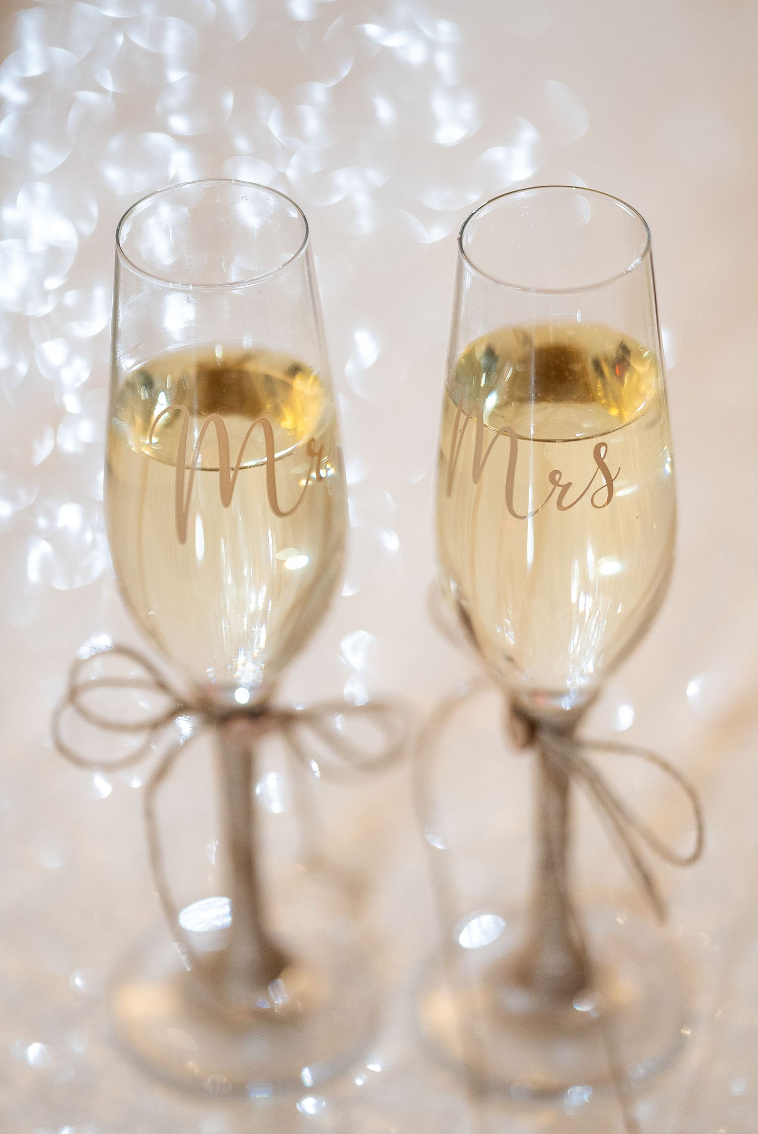 Two champagne flutes with 'Mr & Mrs' text and twine bows filled with sparkling wine against a sparkly background.