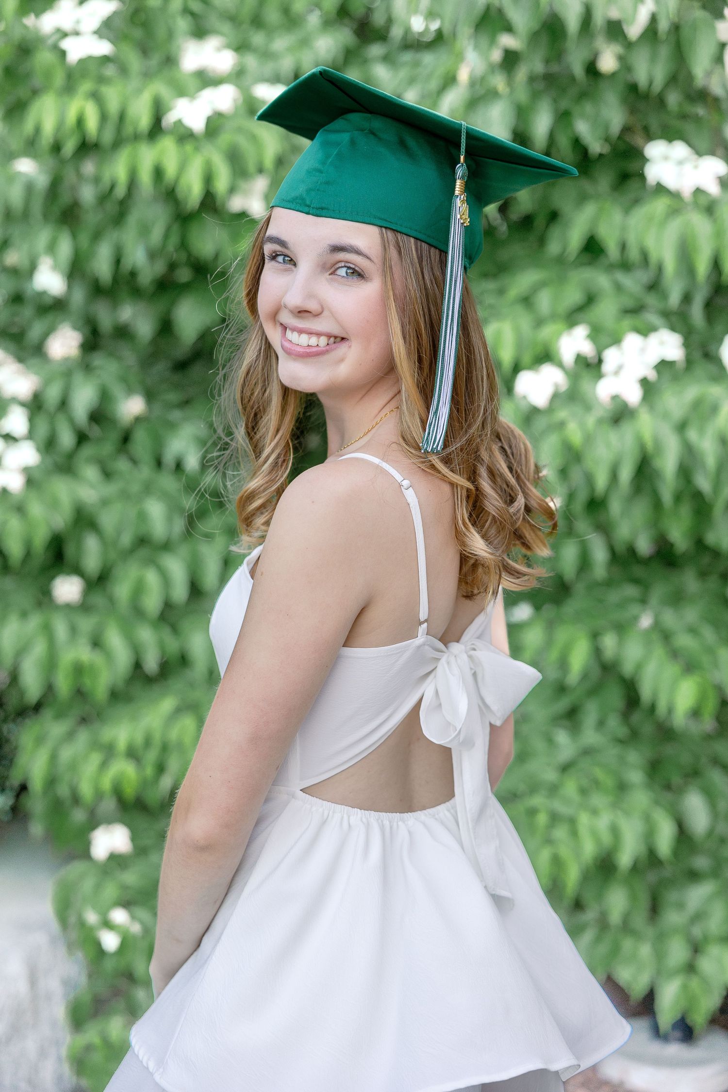 Graduate in white dress and green cap poses outdoors against leafy background.