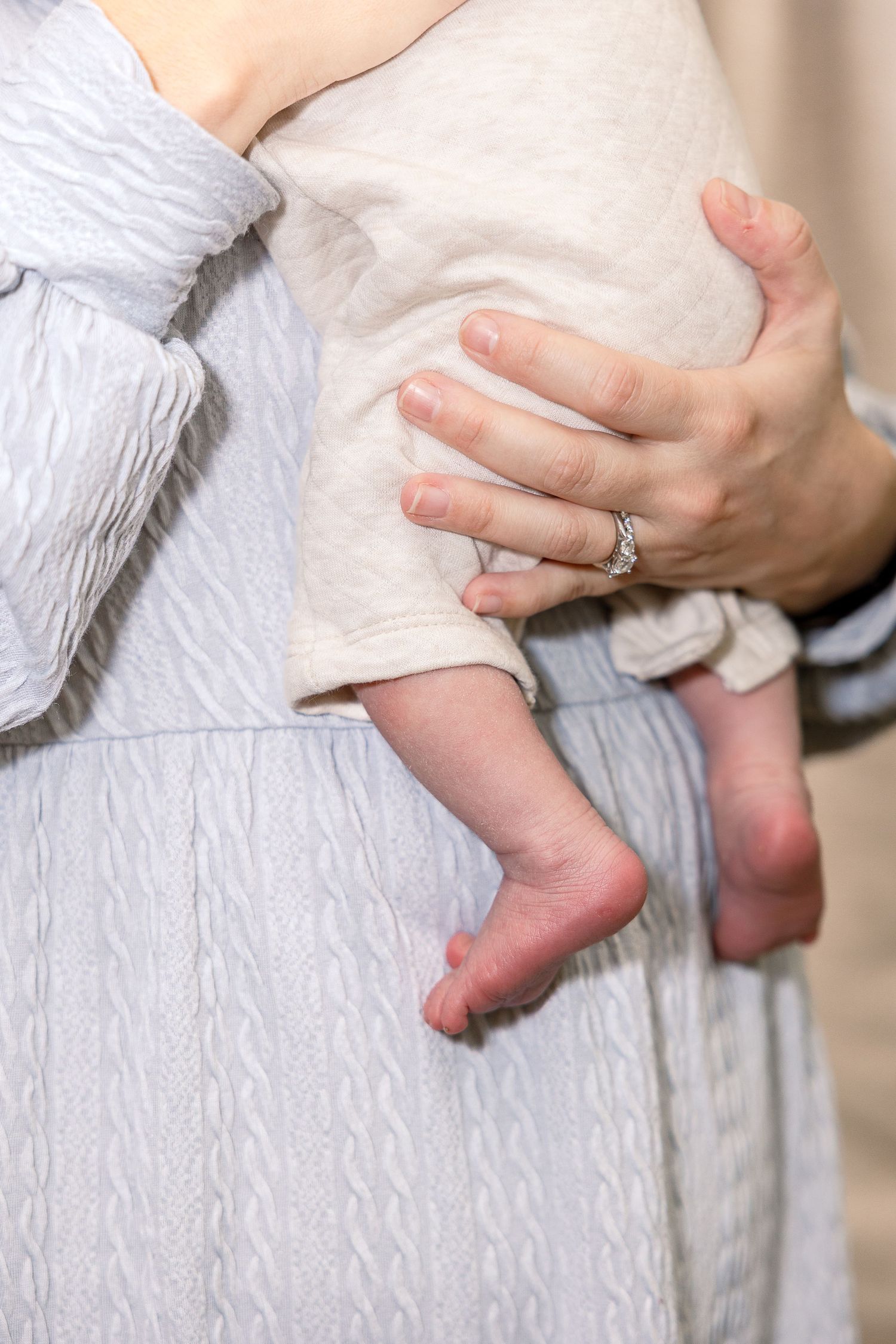 A baby's tiny bare feet dangle next to a woman's hip wearing a light textured dress while a hand with a ring gently holds them.