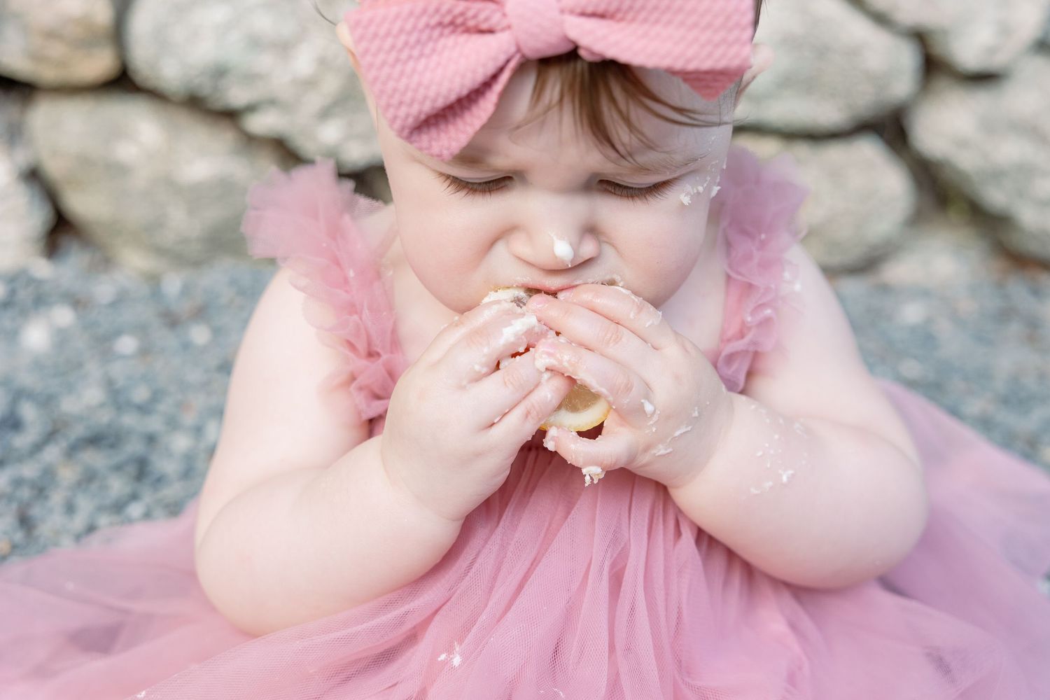 Small figure in pink tulle dress and bow headband takes a big bite of a cookie against stone wall background.