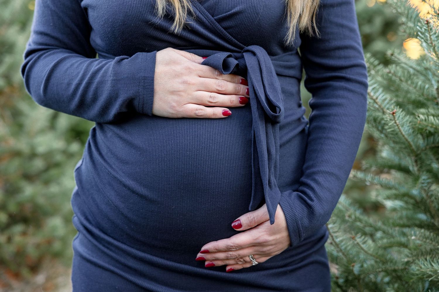 Pregnant woman in navy blue dress cradles her belly while standing near evergreen trees outdoors.