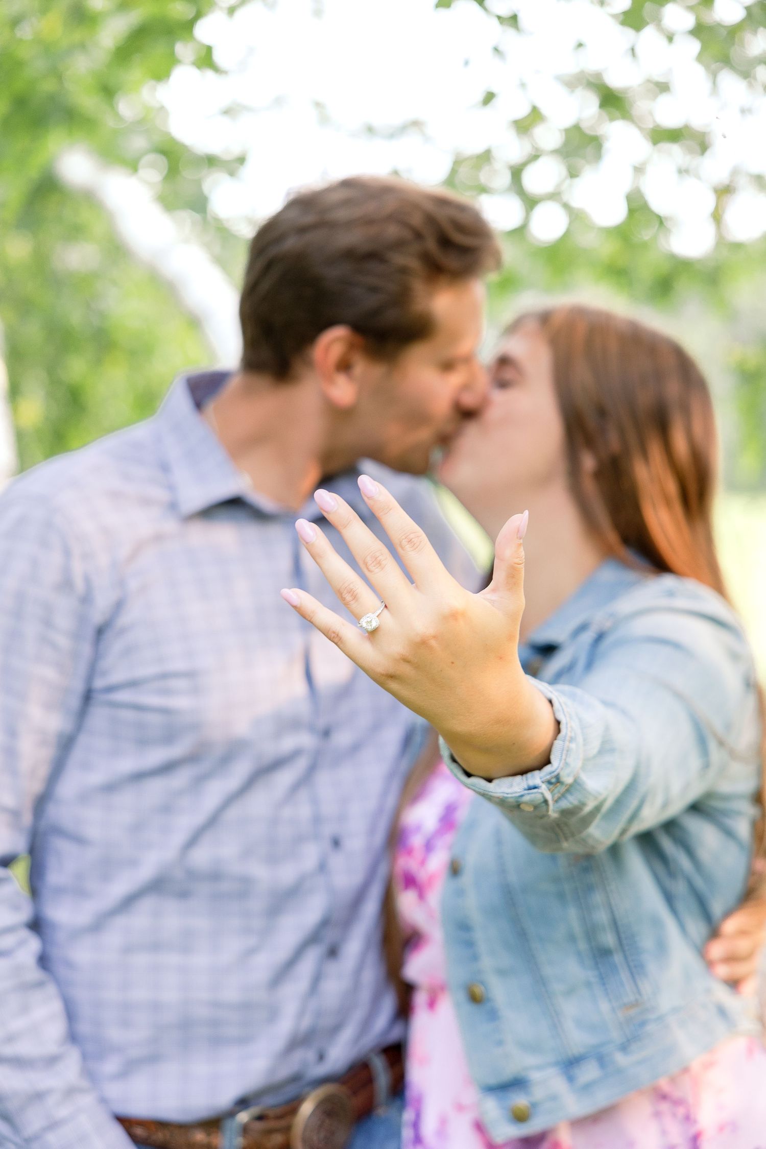 Engaged couple kissing while showing off new ring in outdoor setting.