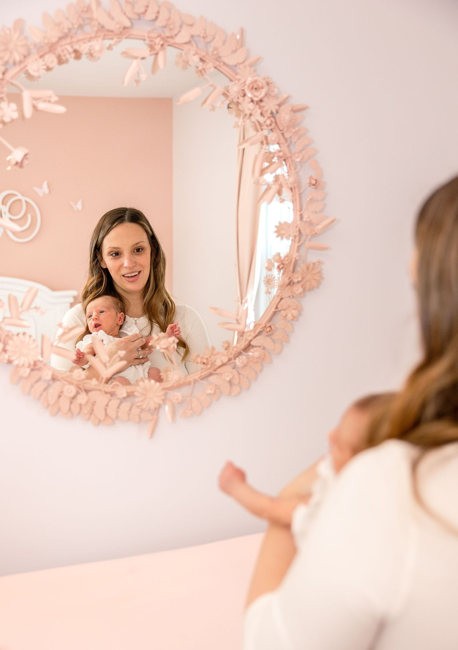 Mirror reflection of someone holding a baby in a nursery with an ornate pink circular frame on a soft pink wall.