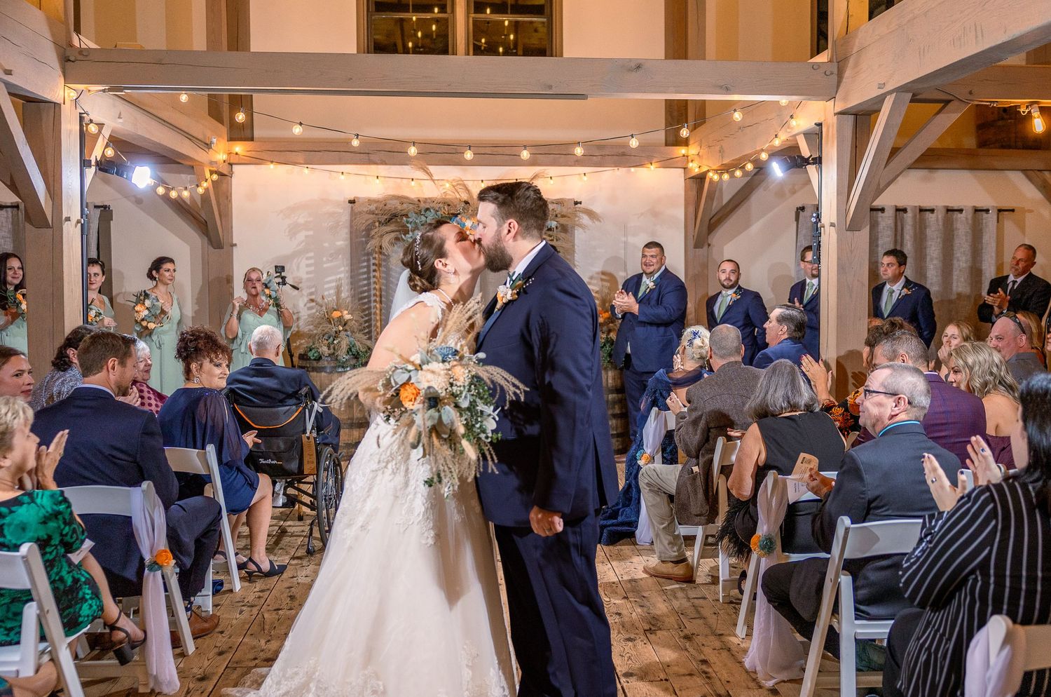 A wedding ceremony takes place in a rustic indoor venue with exposed beams and fairy lights while guests look on.