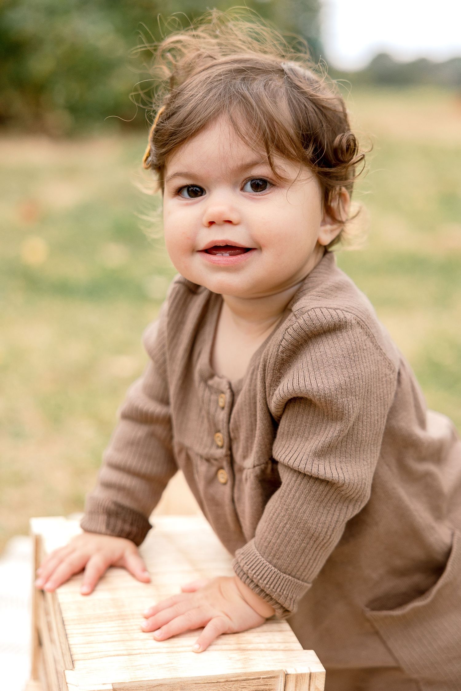 Toddler in brown knit cardigan smiles while leaning on wooden box outdoors.