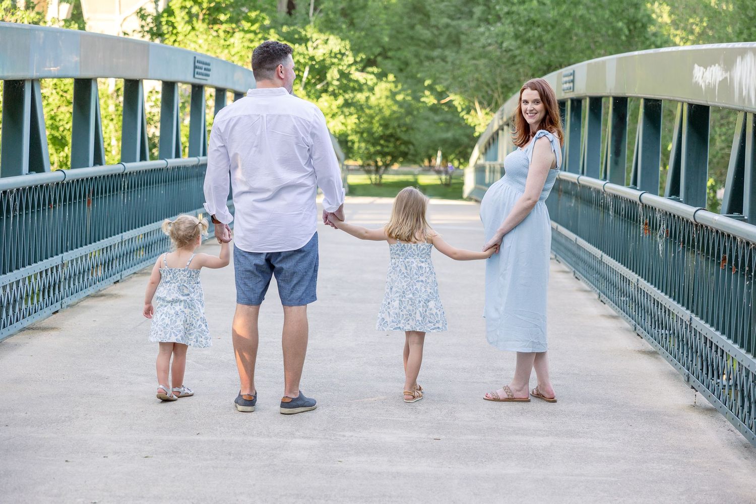 A family holding hands while walking together on a teal-colored bridge in a sunny park setting.