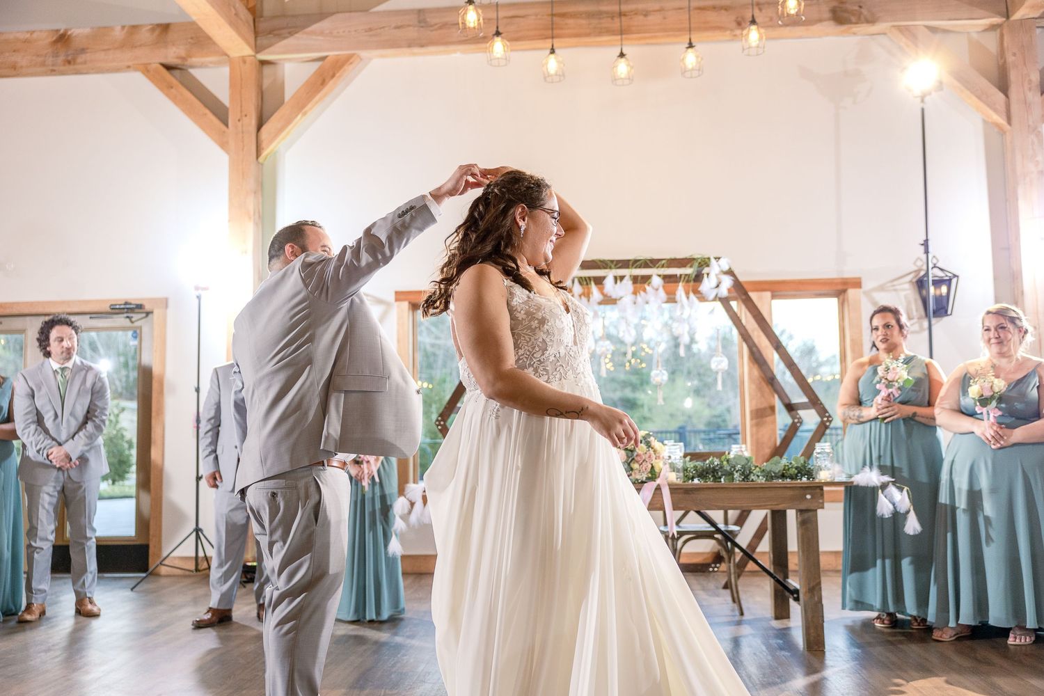 A couple shares their first dance at a rustic barn wedding venue while guests in blue dresses look on.