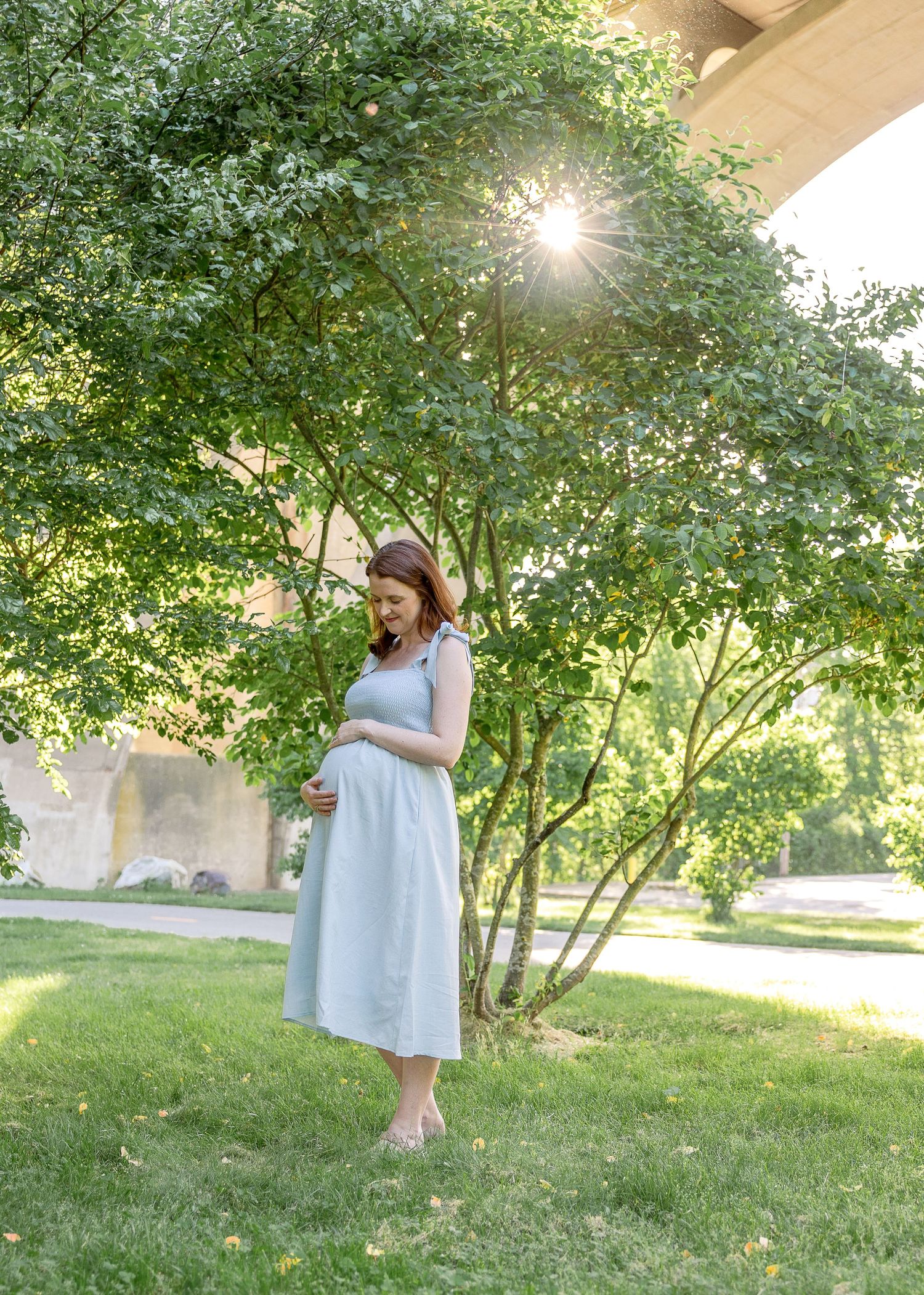 A woman in a light blue dress stands in a sunlit garden setting while cradling her baby bump.