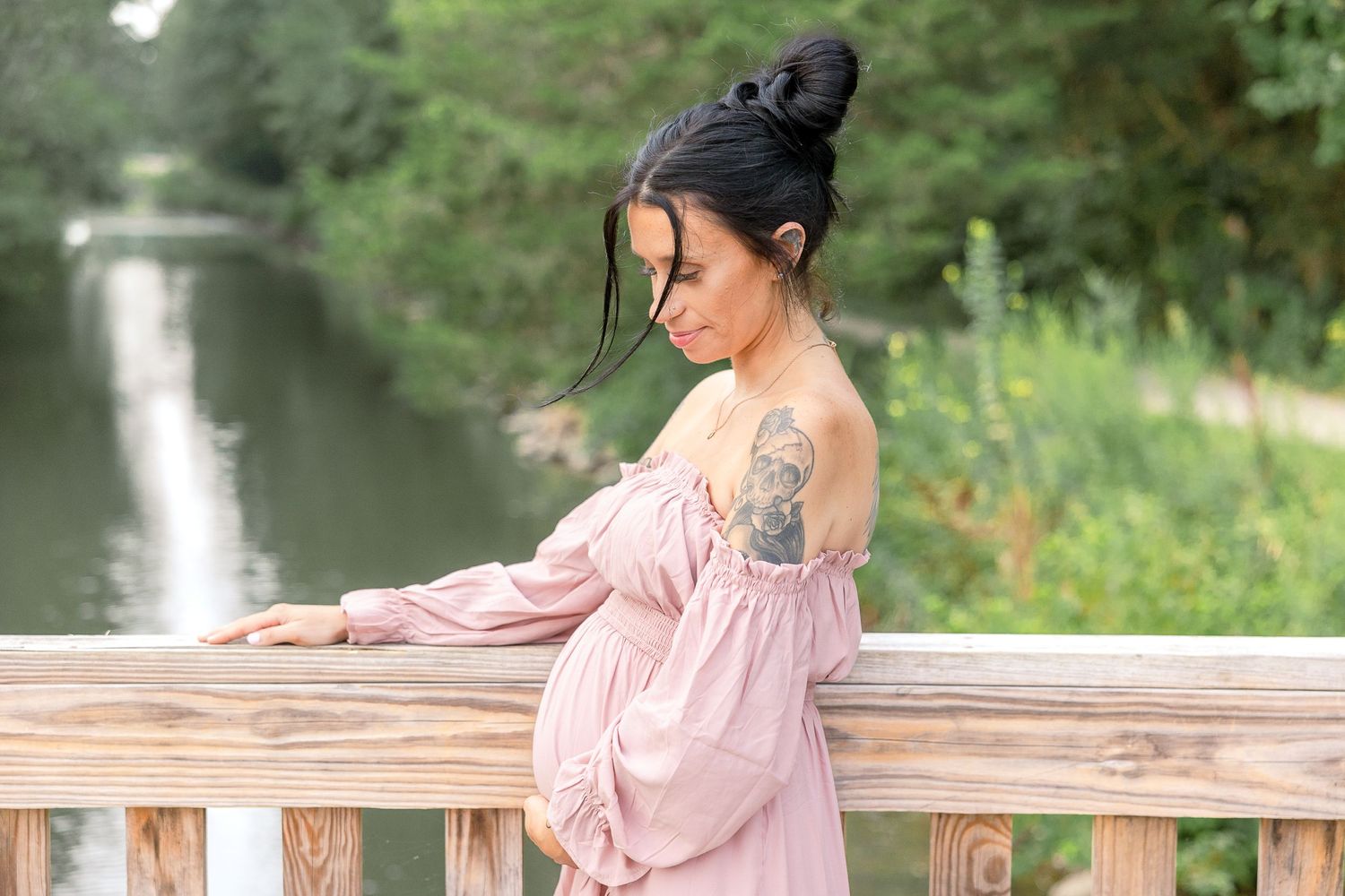 Person wearing pink off-shoulder dress stands at wooden railing along peaceful waterway with green foliage in background.