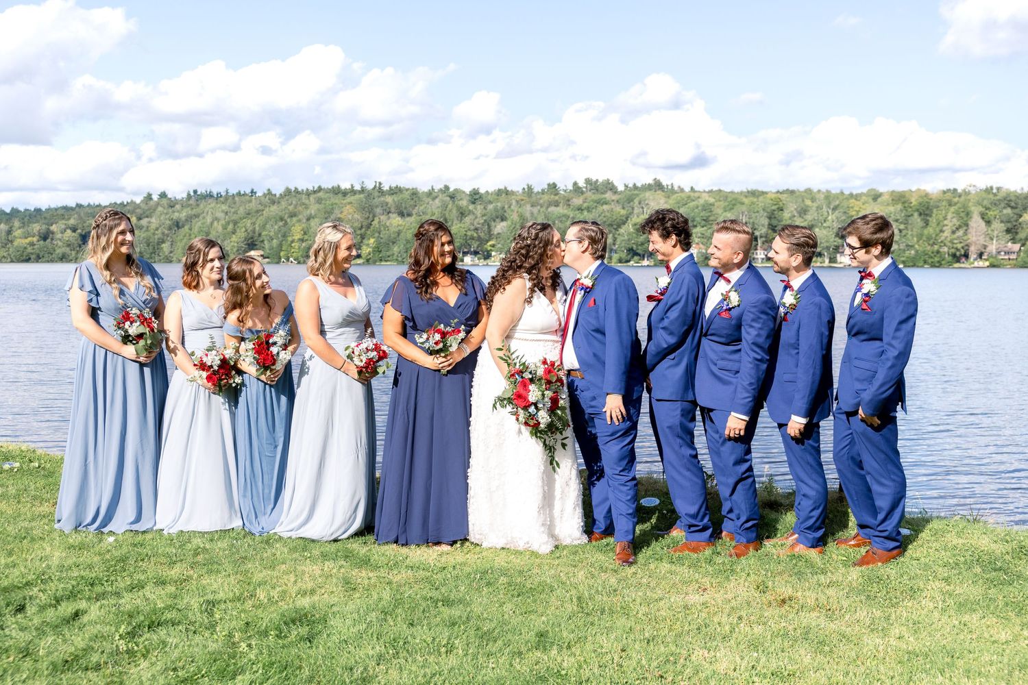 Wedding party in blue and white attire posed together on grass by lakeside.