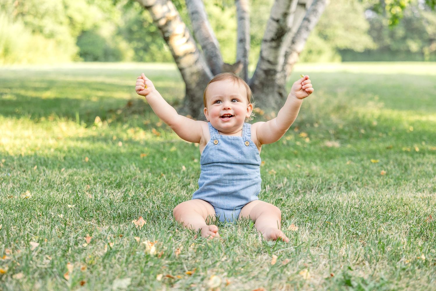 Joyful little one in blue romper sits on grass with arms raised, celebrating in a sunny outdoor setting.