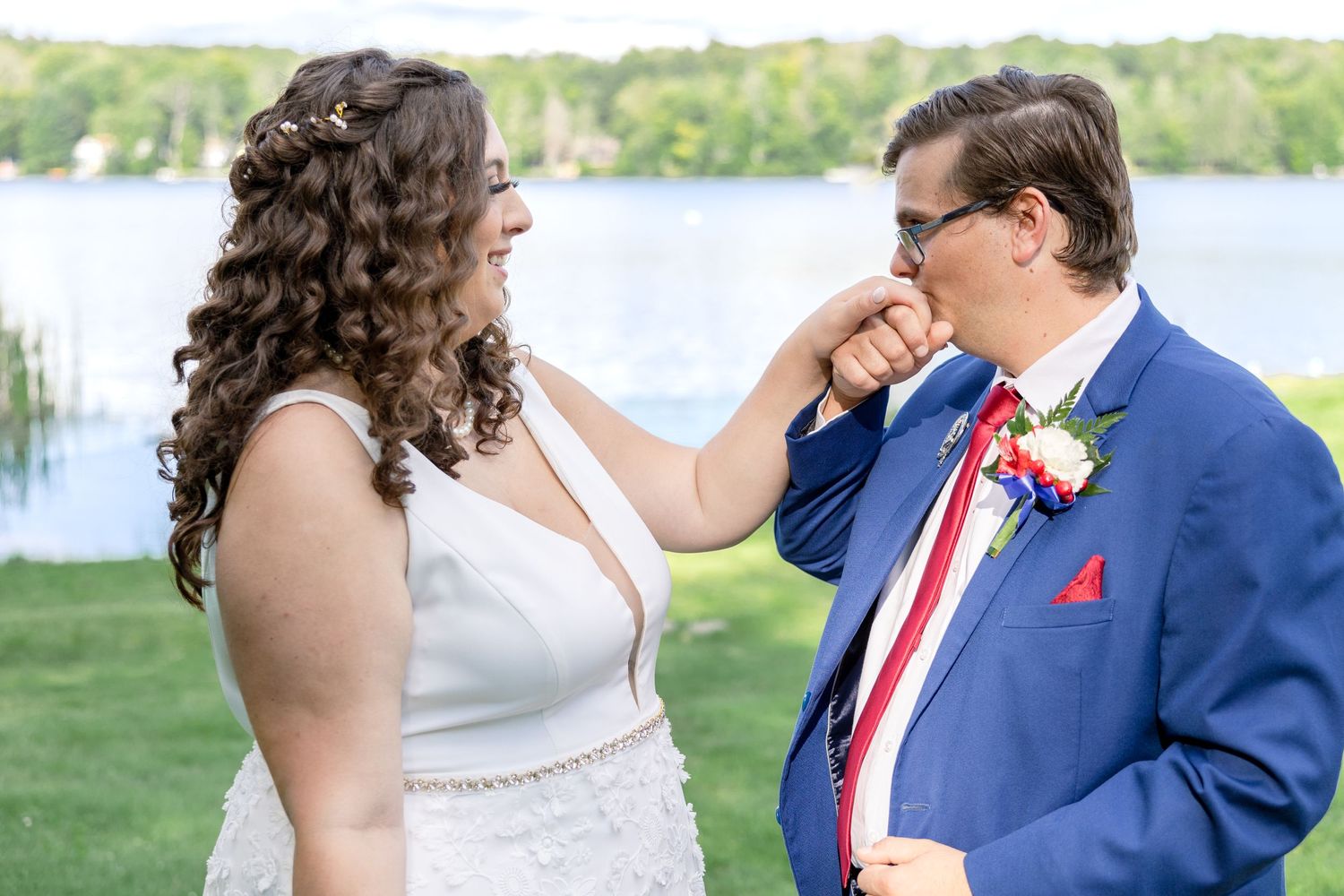 A couple shares a sweet moment during their lakeside wedding ceremony, with the groom in a blue suit and red accessories.