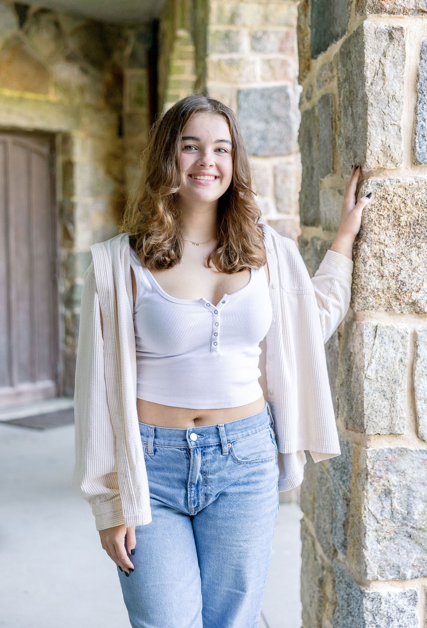 A person in a white crop top and cream cardigan poses against a stone wall in casual denim jeans.