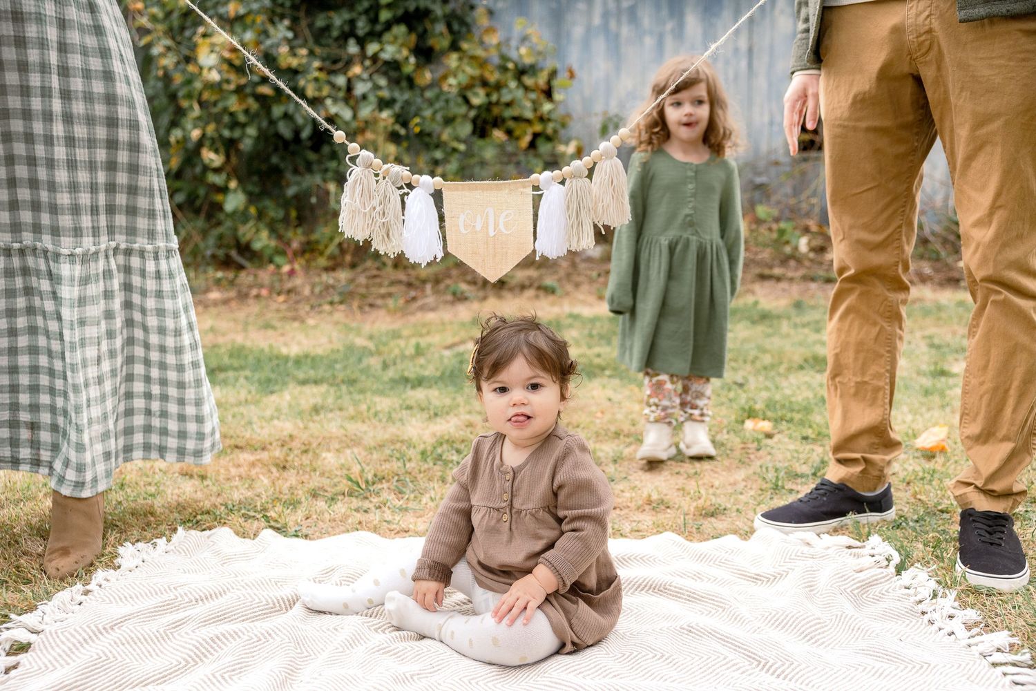 A baby sits on a white blanket in a garden with a clothesline and people standing in the background.