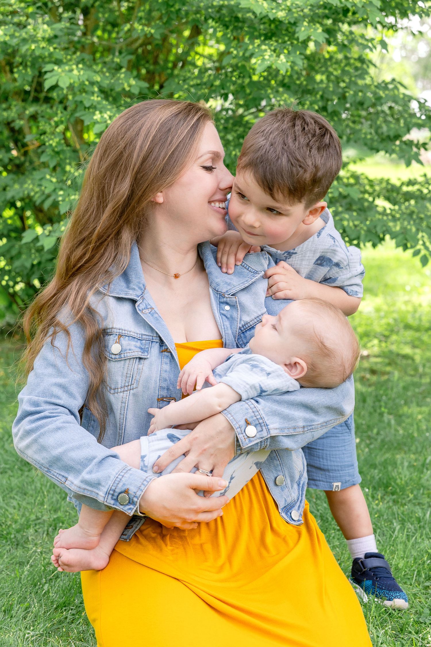Family wearing matching denim jackets enjoys tender moment together on green grass during summer day in park.