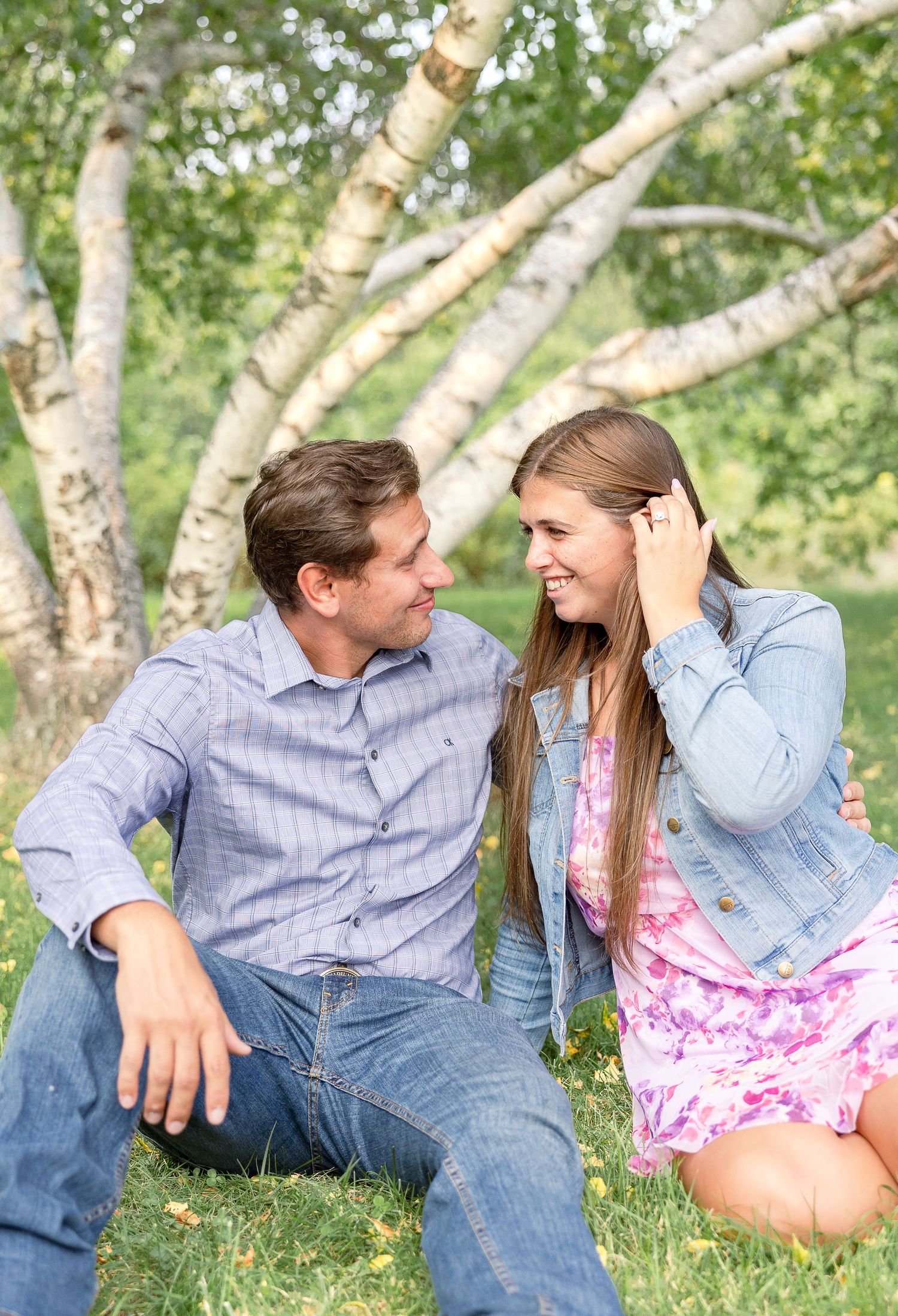 A couple shares a sweet moment together while sitting on grass near birch trees on a sunny spring day.
