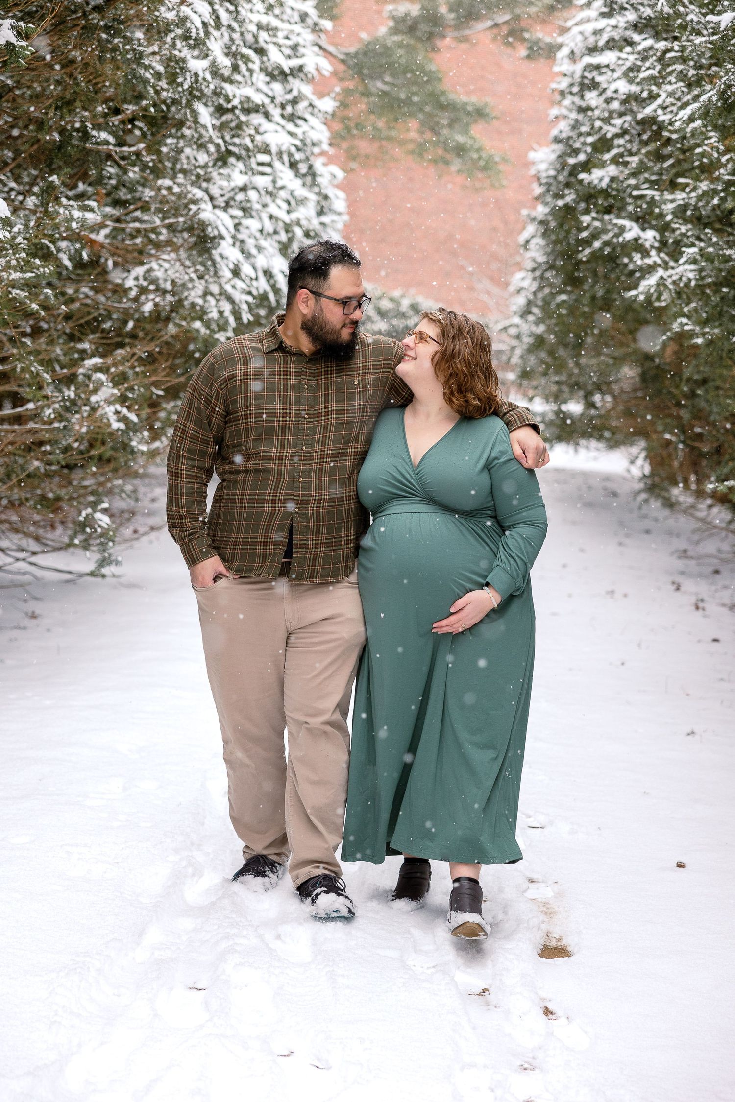 A couple stands together on a snowy path surrounded by snow-covered evergreen trees in a romantic winter scene.