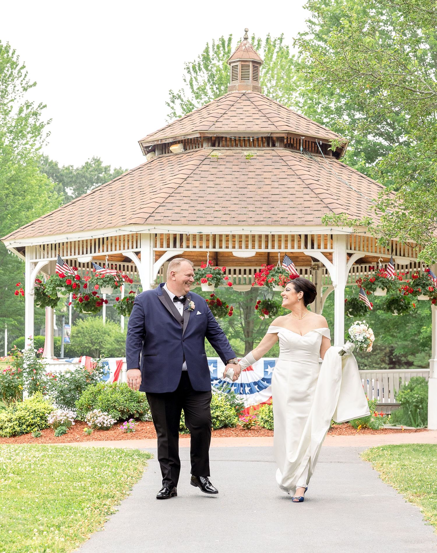 Elegant wooden gazebo with decorative hanging baskets stands in a lush garden setting with blooming flowers and greenery.
