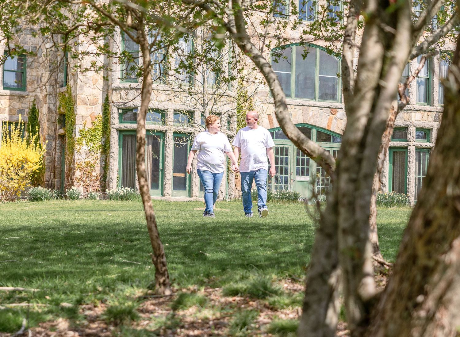 Two people walking together on a green lawn surrounded by trees and buildings on a sunny day.