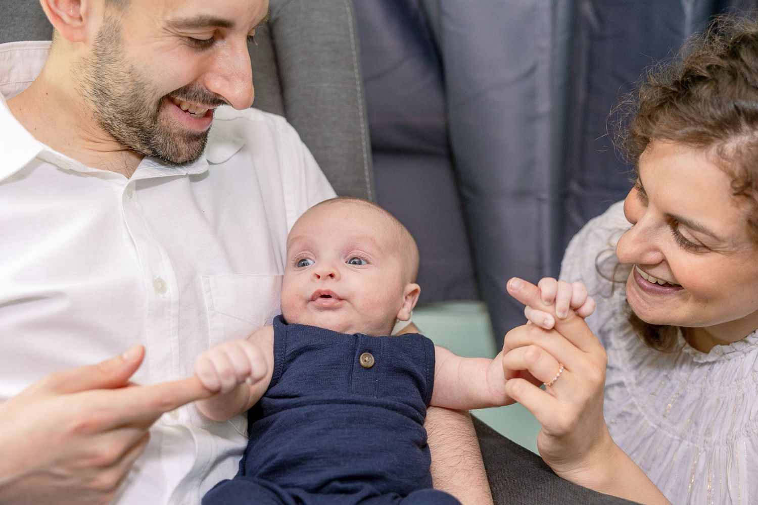Happy parents holding and playing with their smiling baby wearing a navy blue outfit while relaxing at home.