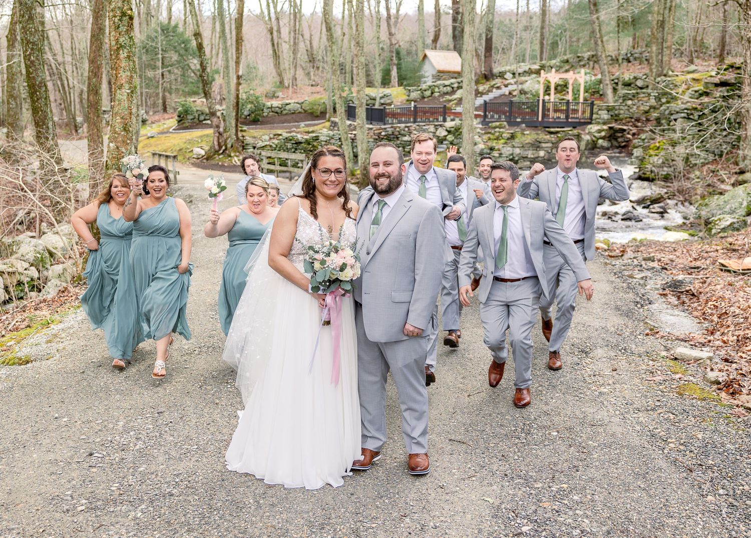 A wedding party walks along a wooded path with a rustic stone wall and autumn foliage in the background.