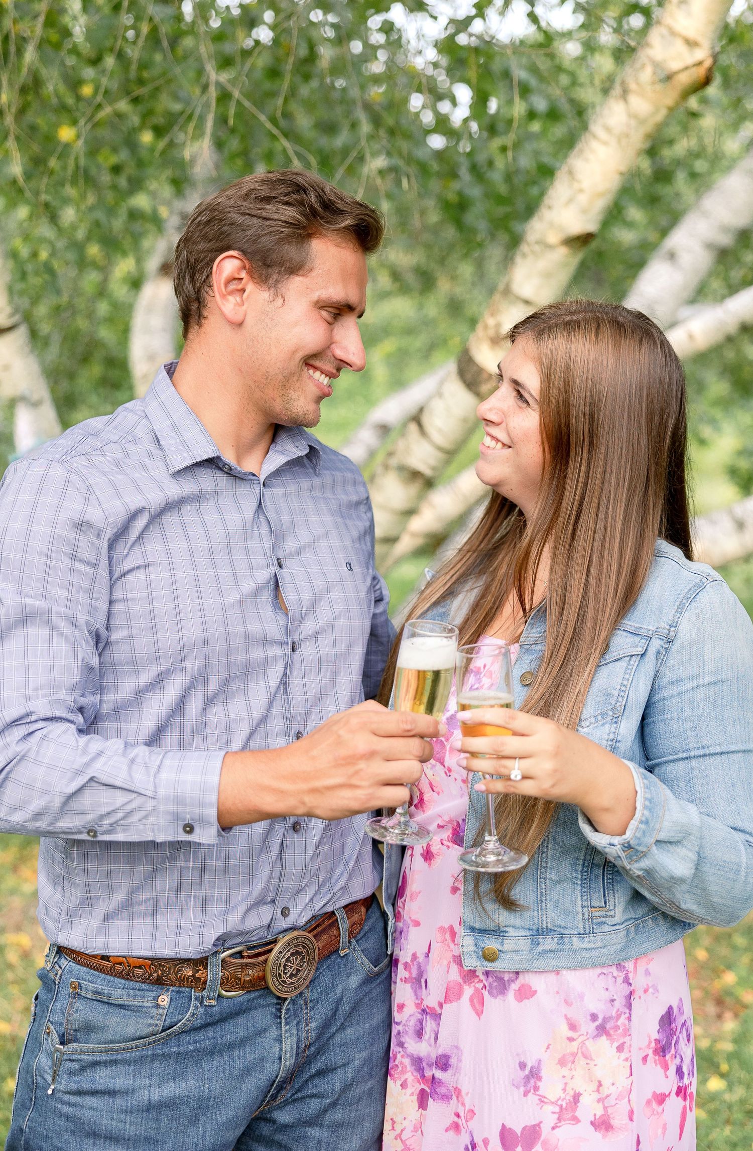 A couple in casual attire share champagne glasses while smiling at each other outdoors near birch trees.