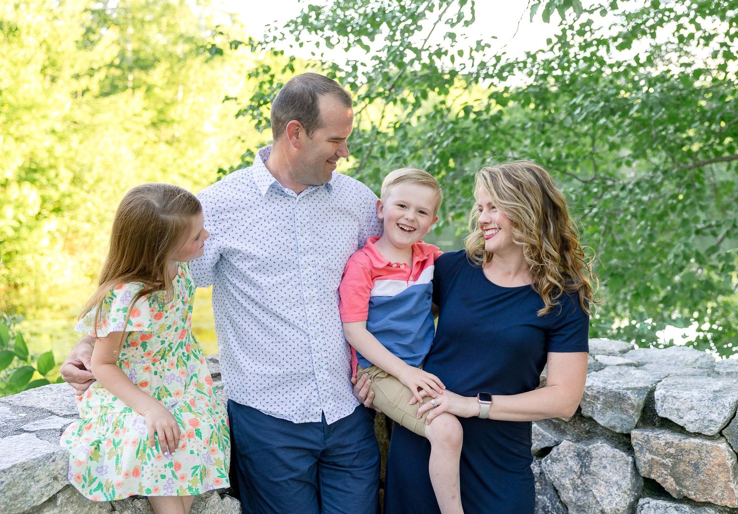 Family shares a candid moment while sitting on stone wall with lush greenery in background.