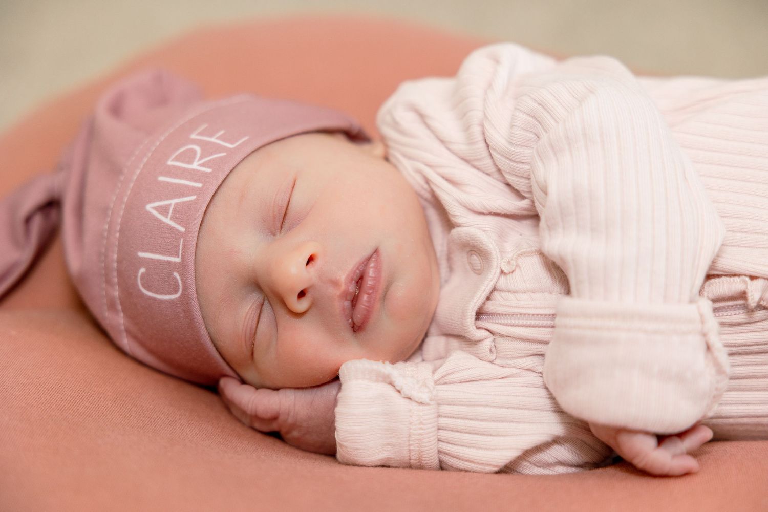 A peaceful sleeping newborn wearing a white knit sweater and personalized beanie rests on a terracotta-colored surface.