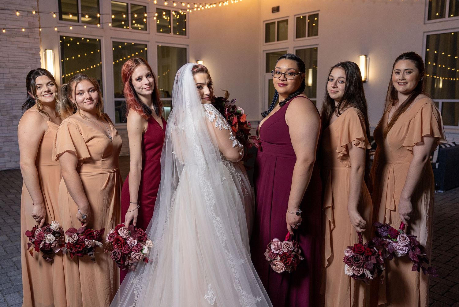 Wedding party of bridesmaids in burgundy and tan dresses holding pink bouquets pose with bride in white gown at reception venue.