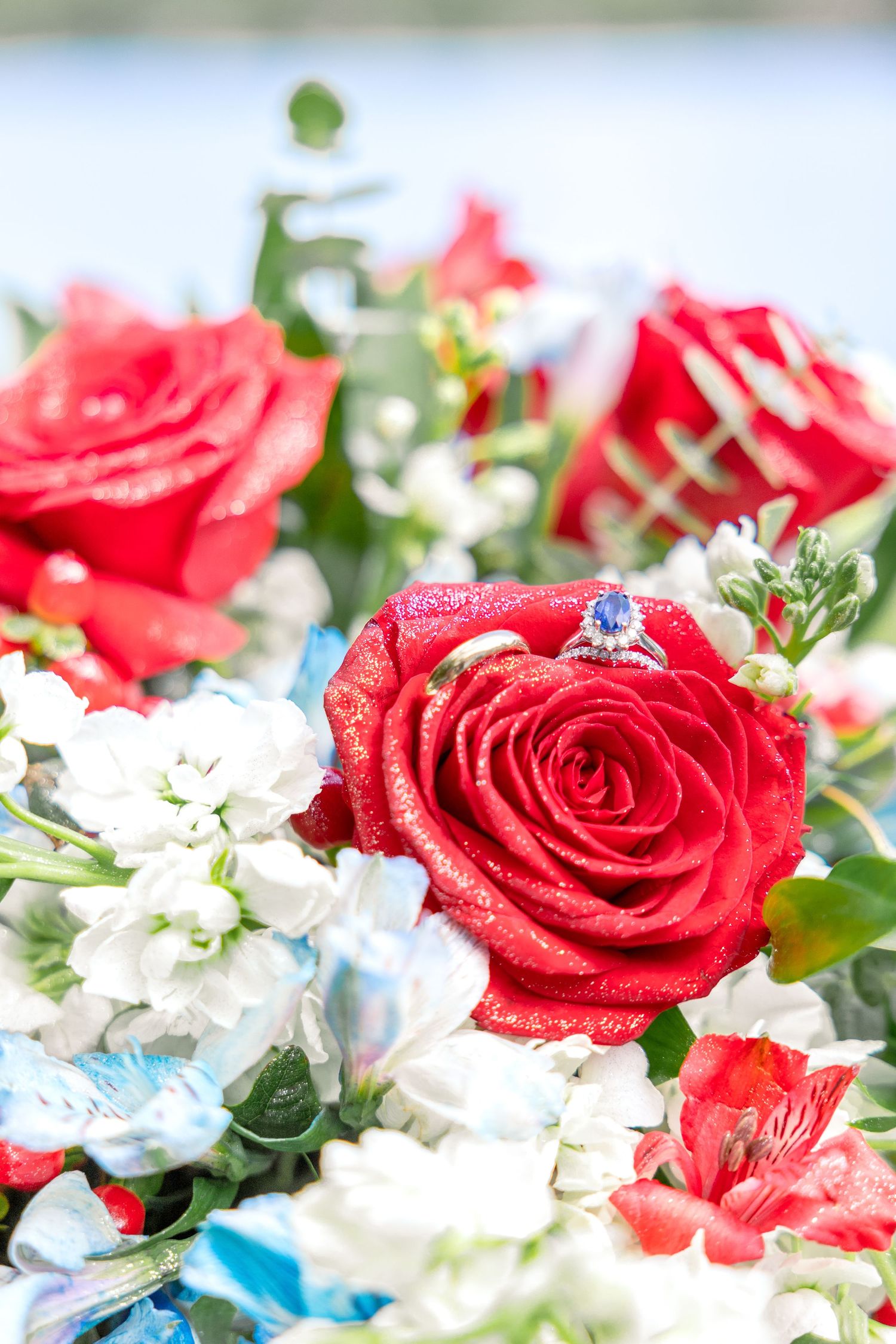 A red rose adorned with a sparkling sapphire ring nestled among white flowers and blue floral accents.