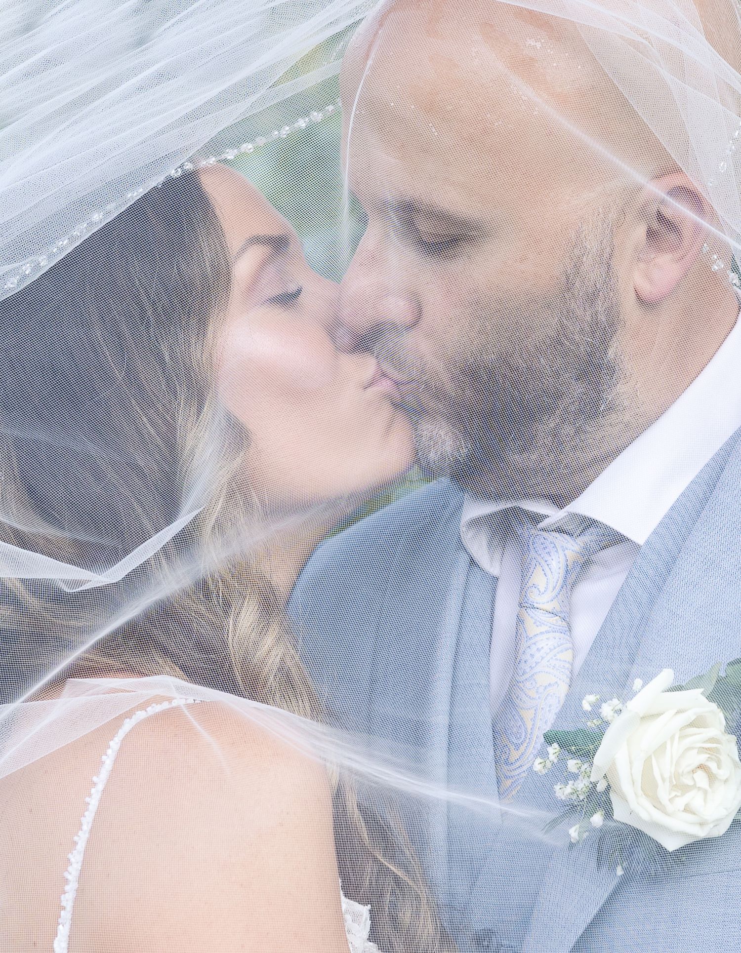 A tender wedding kiss between newlyweds under a flowing white veil with a white rose boutonniere.