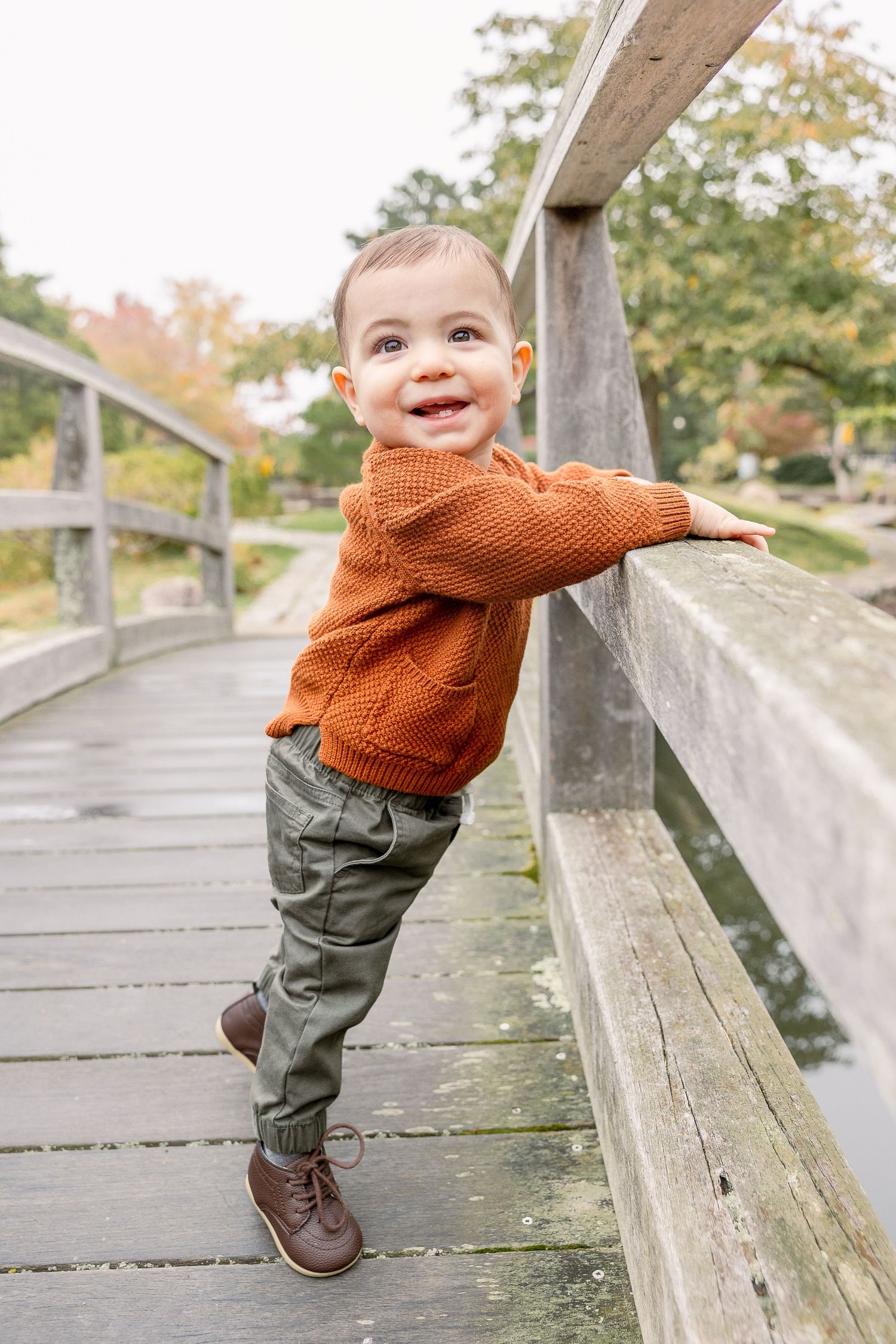 Young child in orange sweater and brown boots leaning against wooden bridge railing during autumn.