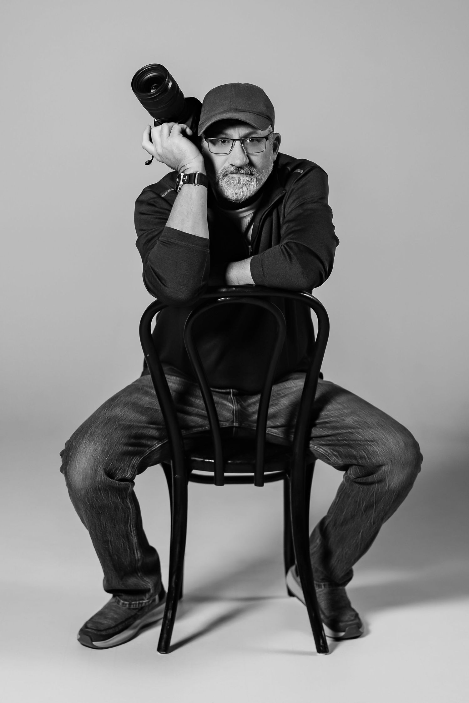 Black and white portrait of a person sitting backwards on a wooden chair while holding a camera in casual attire.