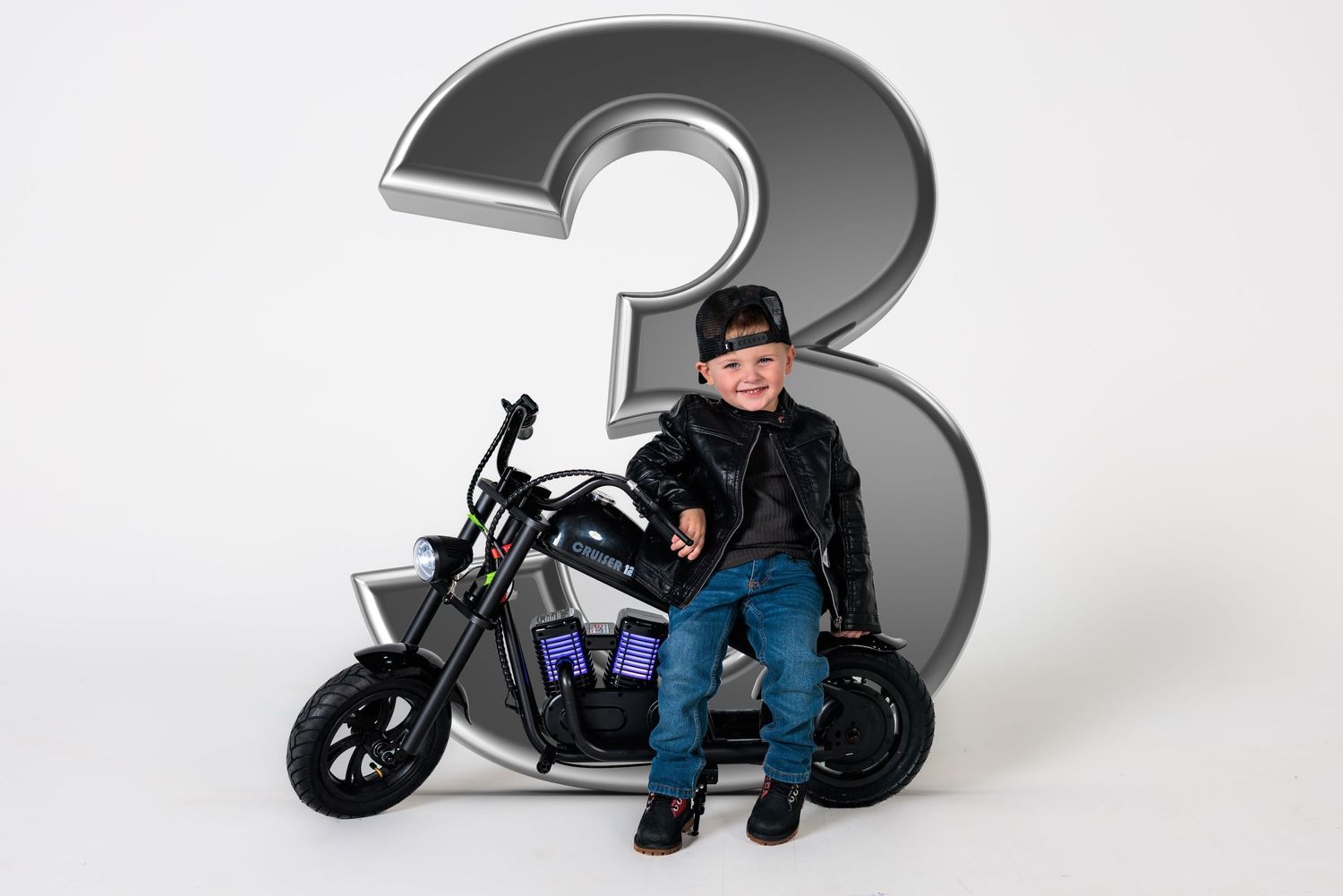 A person sits on a customized motorcycle in front of a large grey number 3 against a white background.