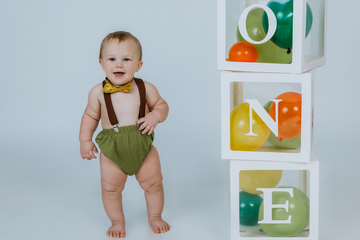Baby wearing brown and green suspender outfit stands next to white cubes with colorful letters.