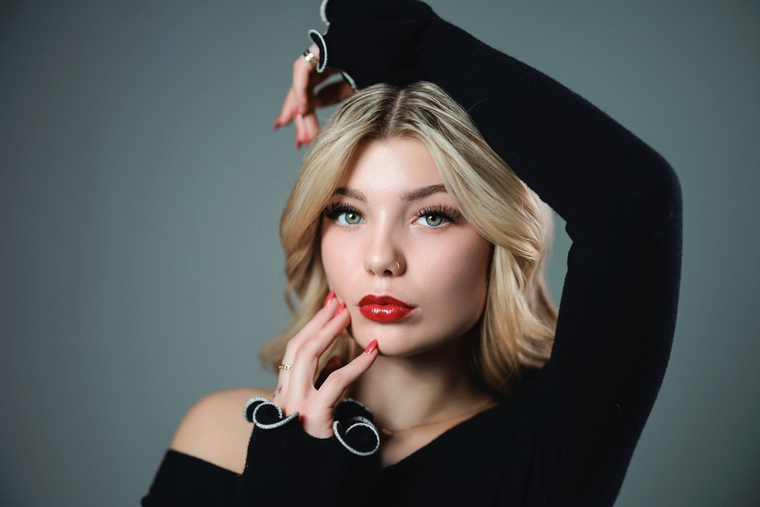 Glamorous beauty portrait featuring bold red lipstick, blonde hair, and a black sweater against gray background.