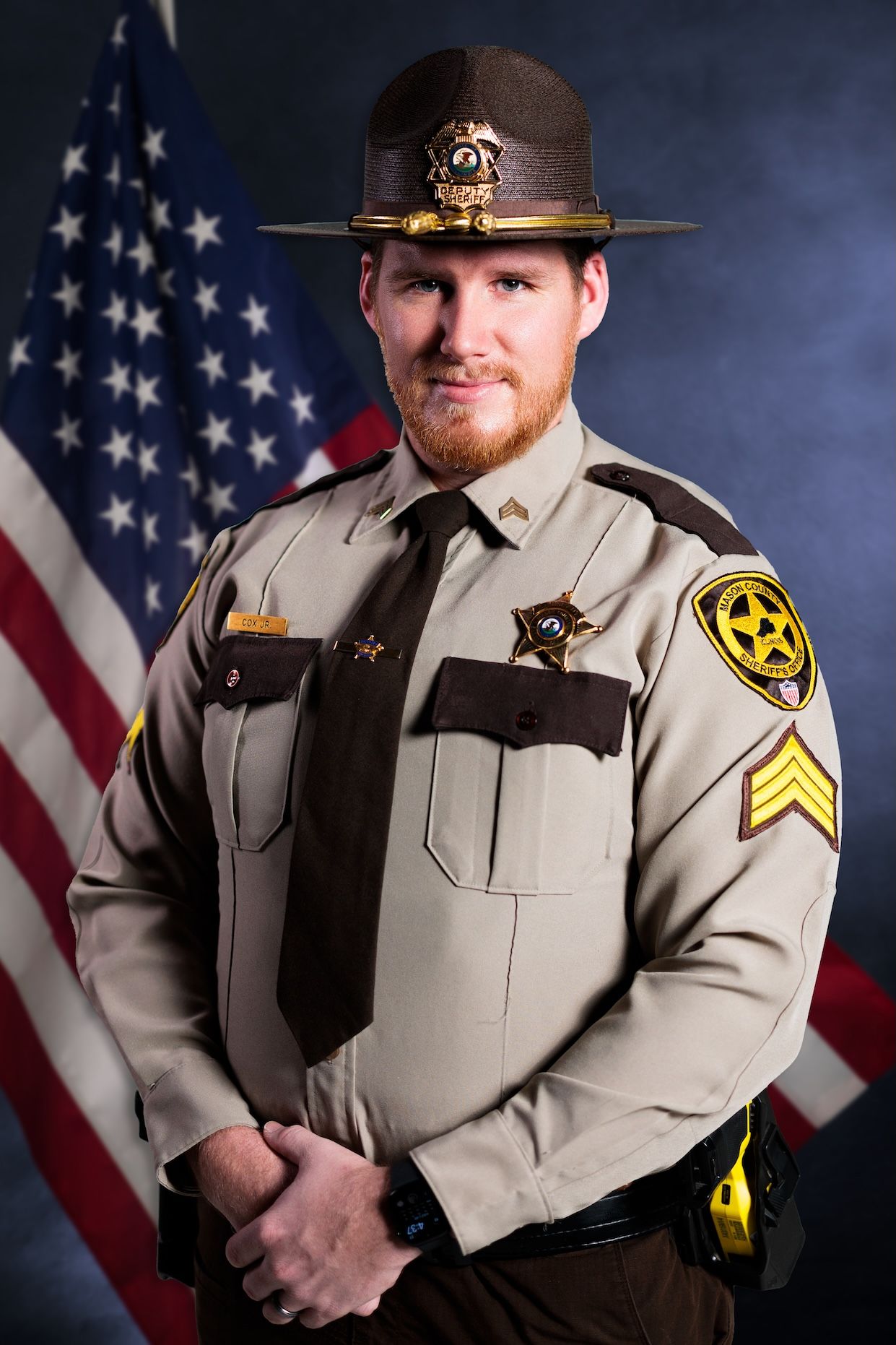 A law enforcement officer in tan uniform poses in front of an American flag for an official portrait.