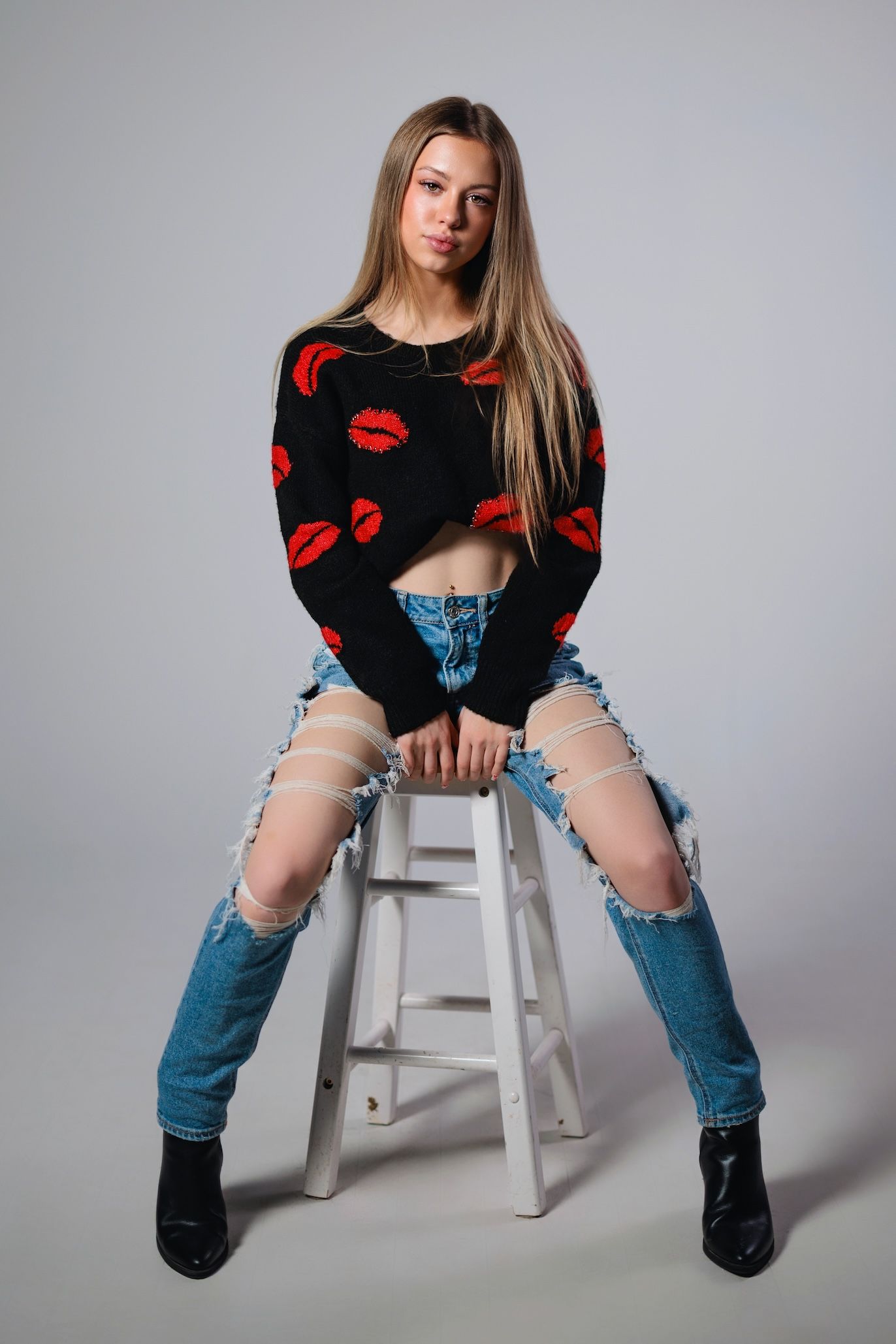 Model in ripped jeans and black sweater with red kiss prints poses on white stool against gray background.