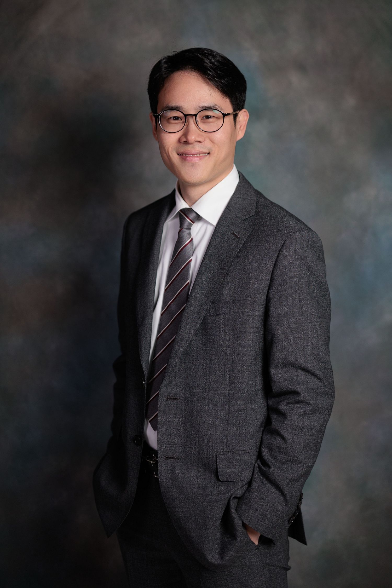 Professional portrait of someone wearing glasses, a gray suit and striped tie against a mottled dark background.