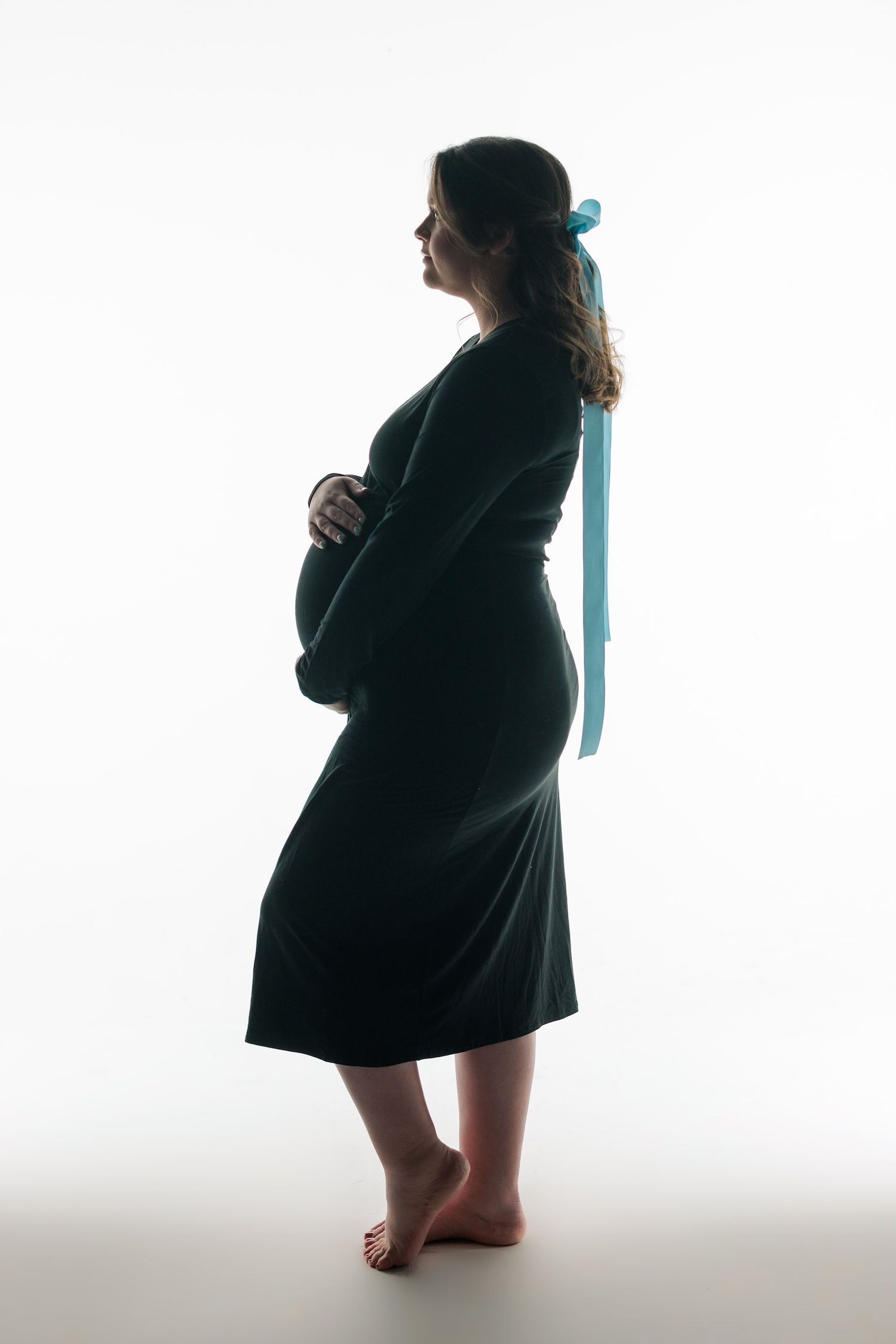 Silhouette of pregnant woman in black dress with blue ribbon standing barefoot against white background.