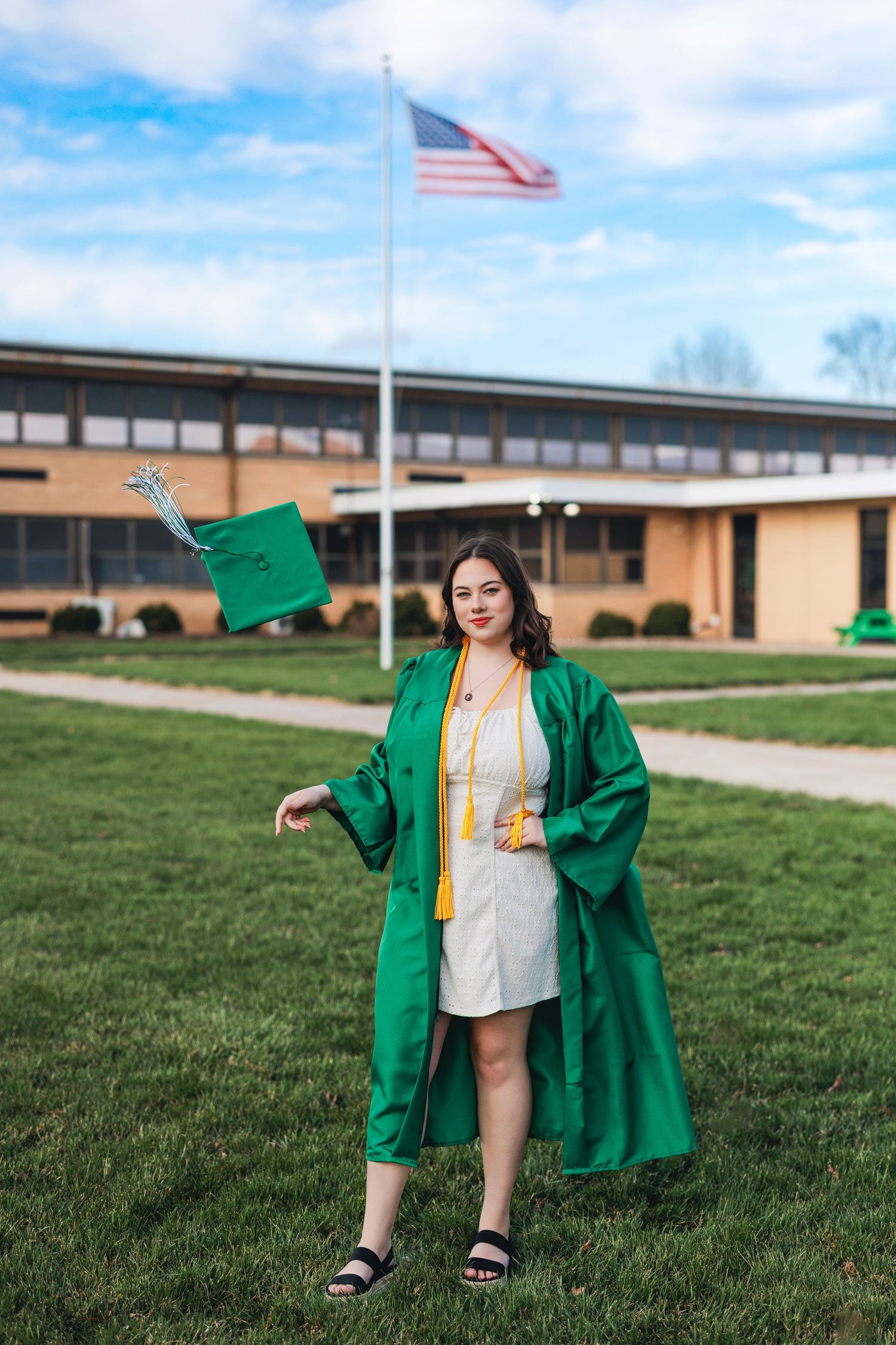 Graduate in emerald green cap and gown poses on campus lawn with American flag flying in background.
